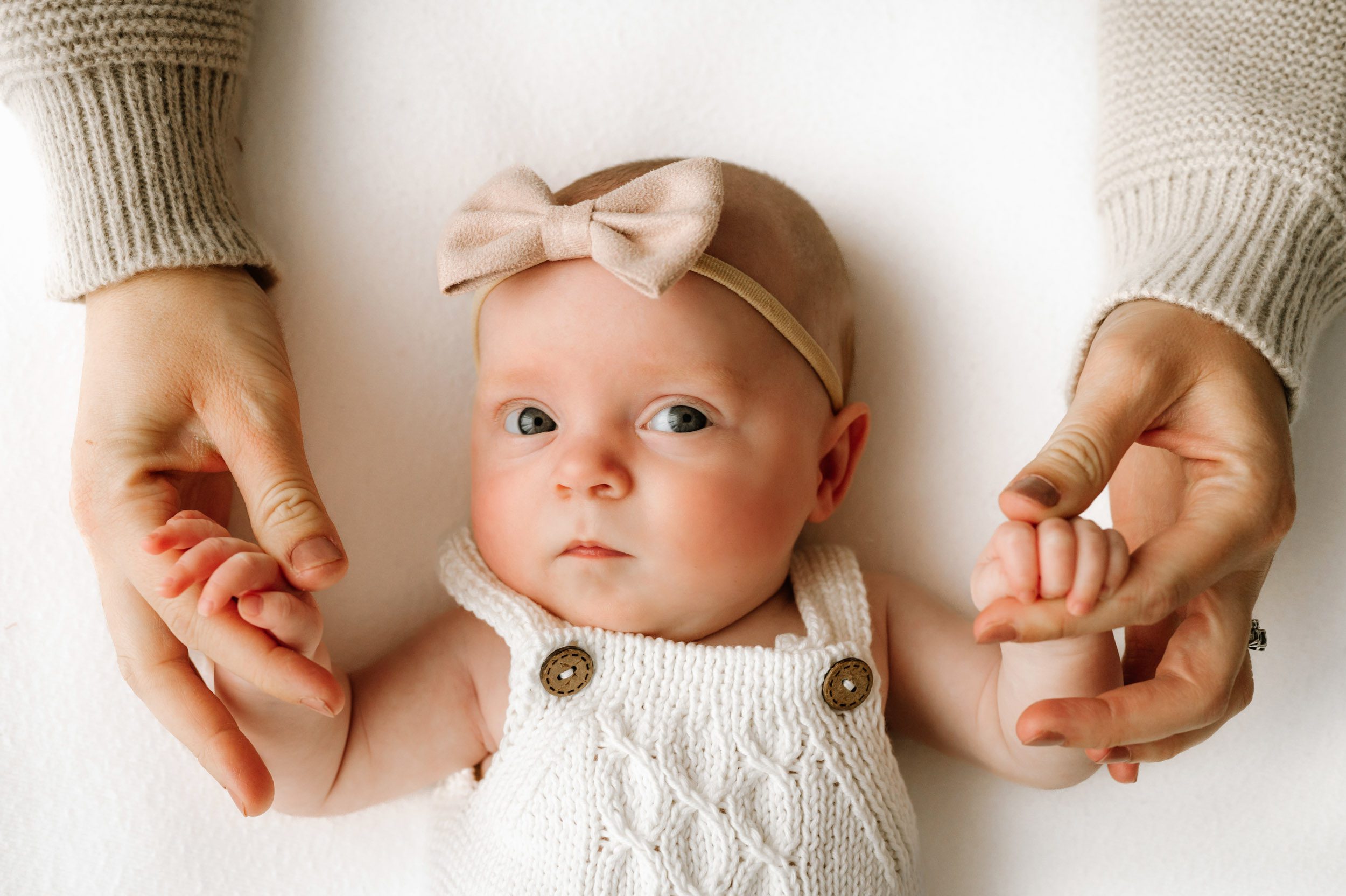 a baby girl wearing a white textured knit romper and a light pink headband holding onto her mom's fingers with each hand and looking up toward the camera during a newborn photo session