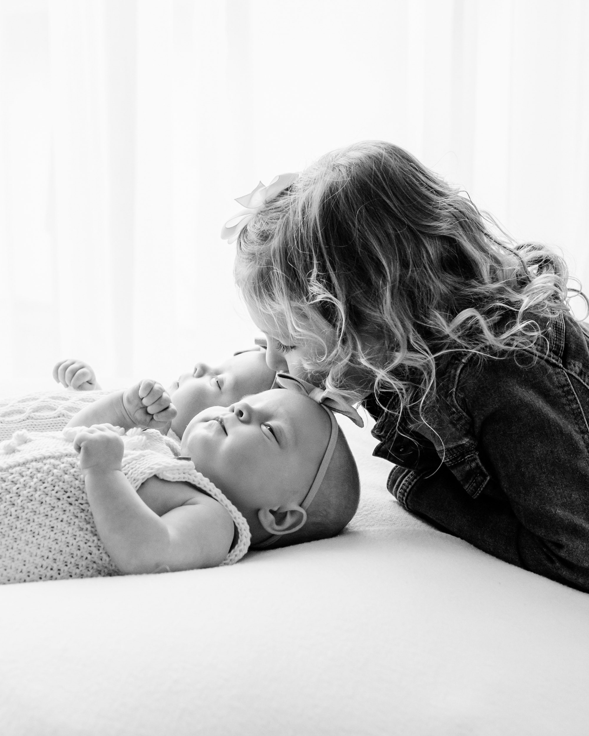 a black and white backlit photo of twins laying on a white beanbag as their older sister kisses them on the head during a lifestyle newborn photoshoot