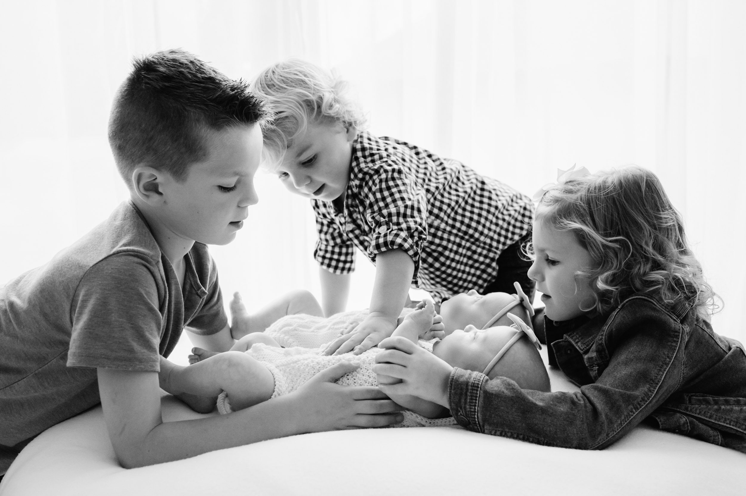 a black and white backlit photo of twins laying on a white beanbag as their two older brothers and older sister all touch the babies with their hands during a lifestyle newborn photoshoot