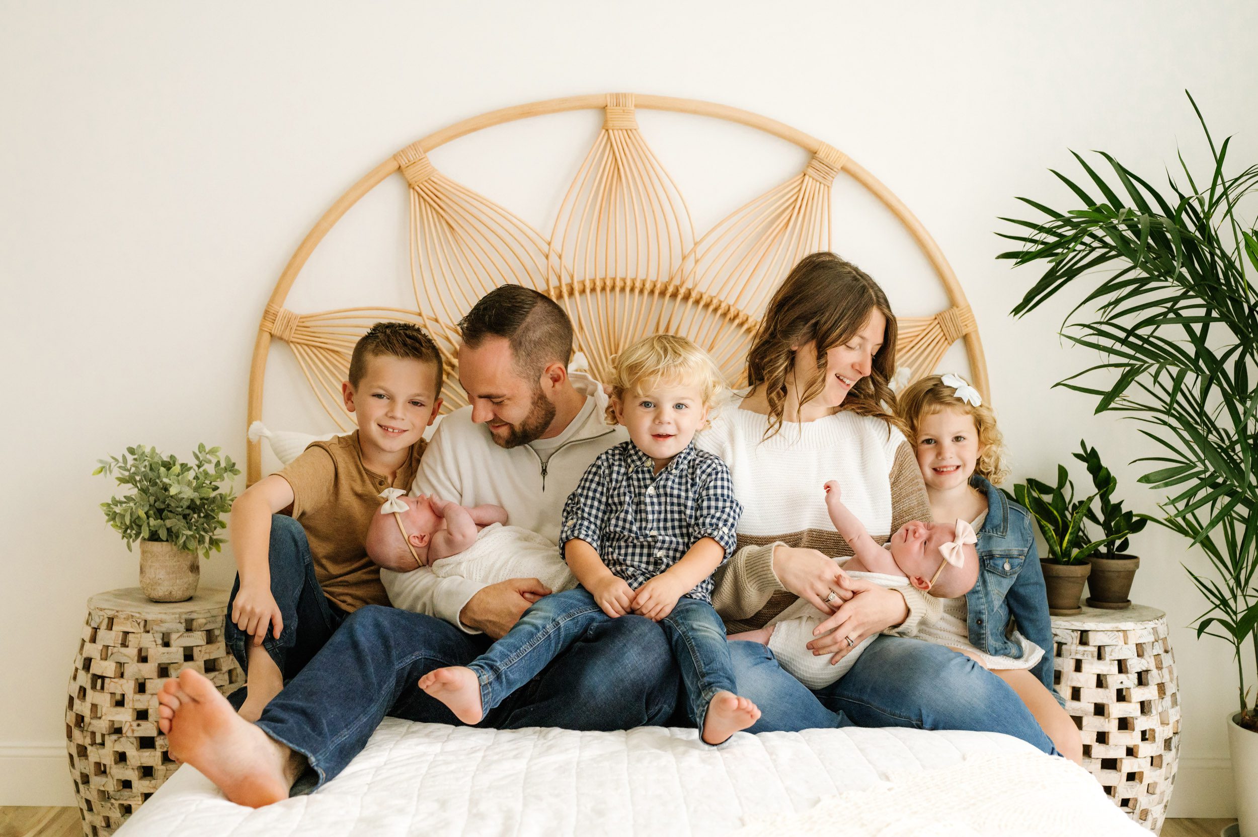 a family of seven sitting on a bed snuggling while mom and dad each cradle one of their twin baby girls in their arms and smile down at them and the three older kids smile directly at the camera during a lifestyle newborn photoshoot