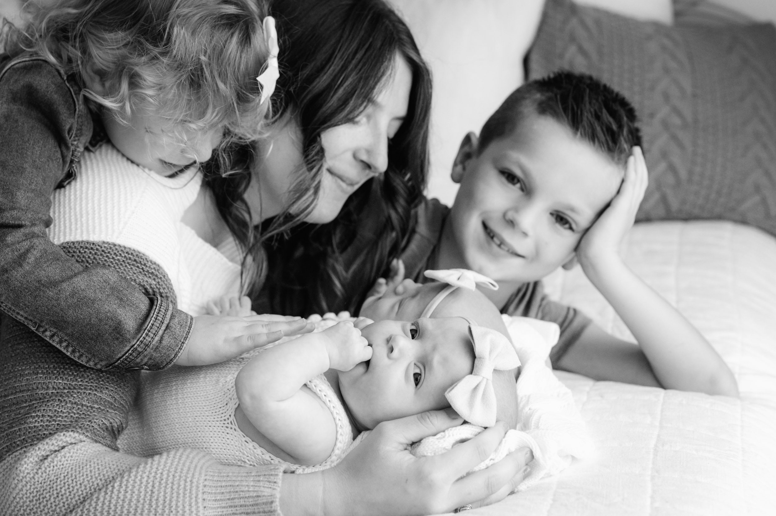 a black and white picture of a mom laying on a bed snuggling with her twin baby girls and two older kids as one of the baby girls looks right at the camera and sucks on her finger during a lifestyle newborn photos session with twins