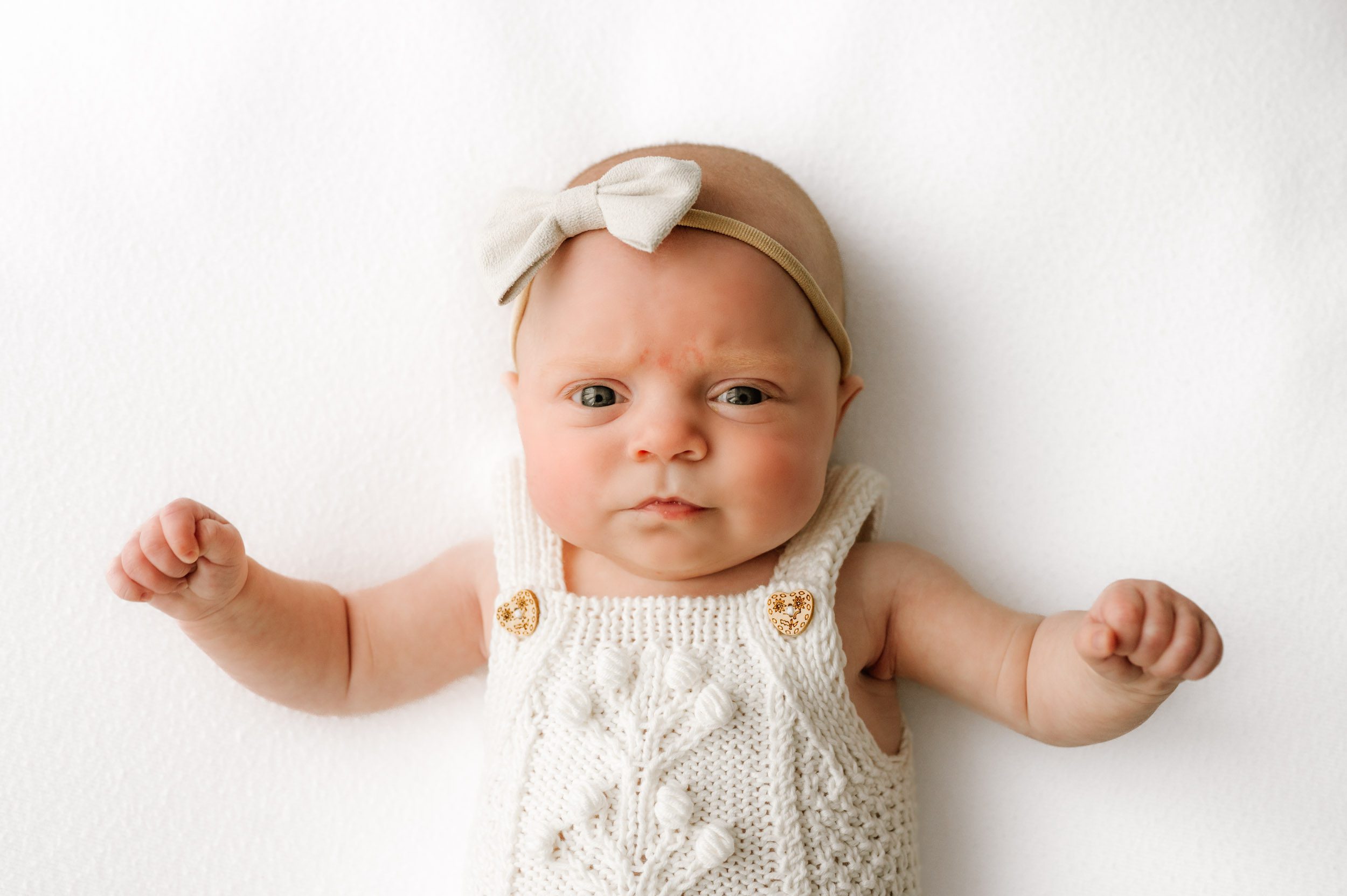 a baby girl wearing a white textured knit romper laying on her back on a white backdrop and looking right at the camera with a concerned look on her face during a newborn photo session