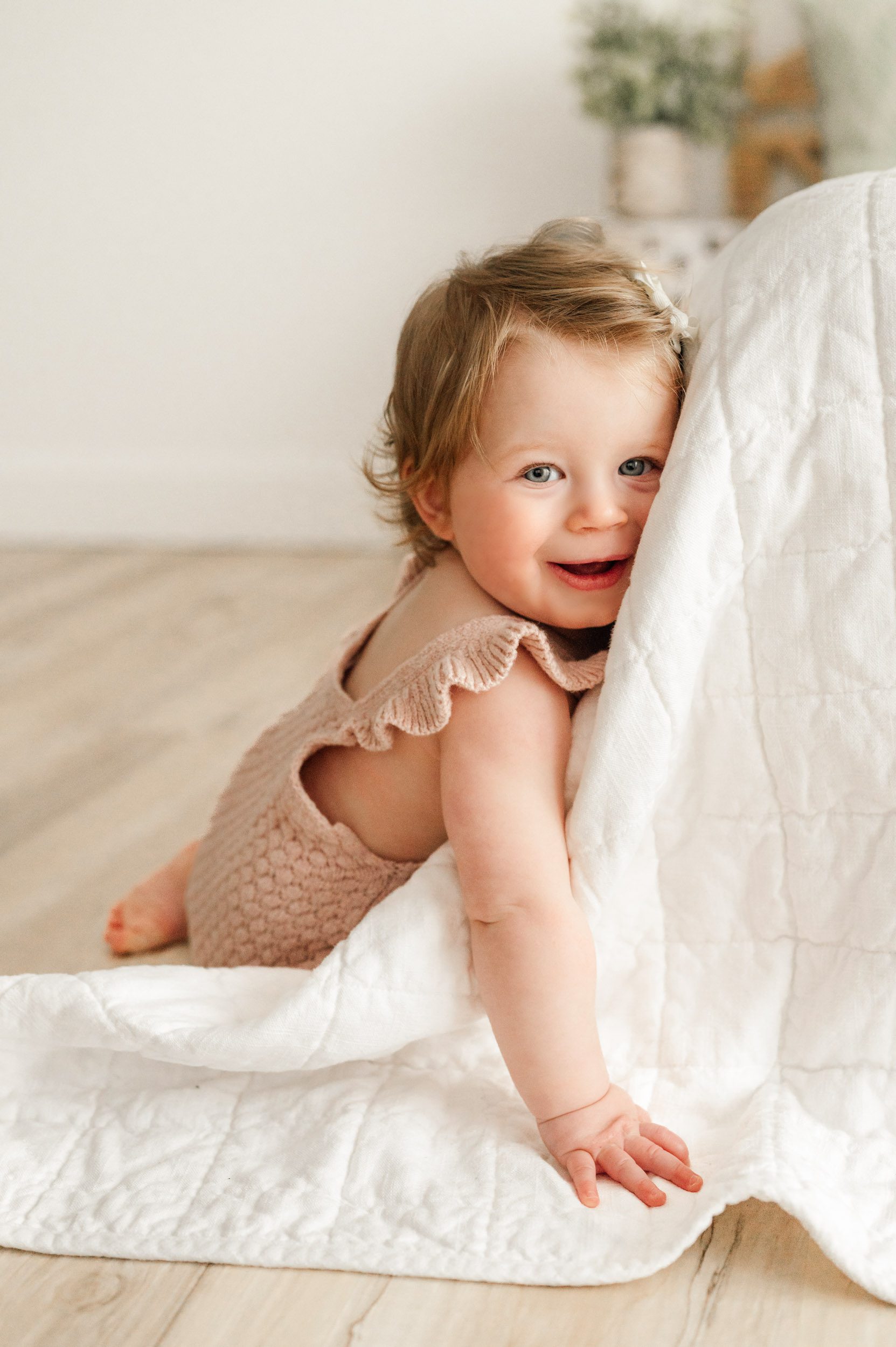 a little girl resting her head against the corner of a bed and smiling at the camera during a Pottstown baby milestone photography session