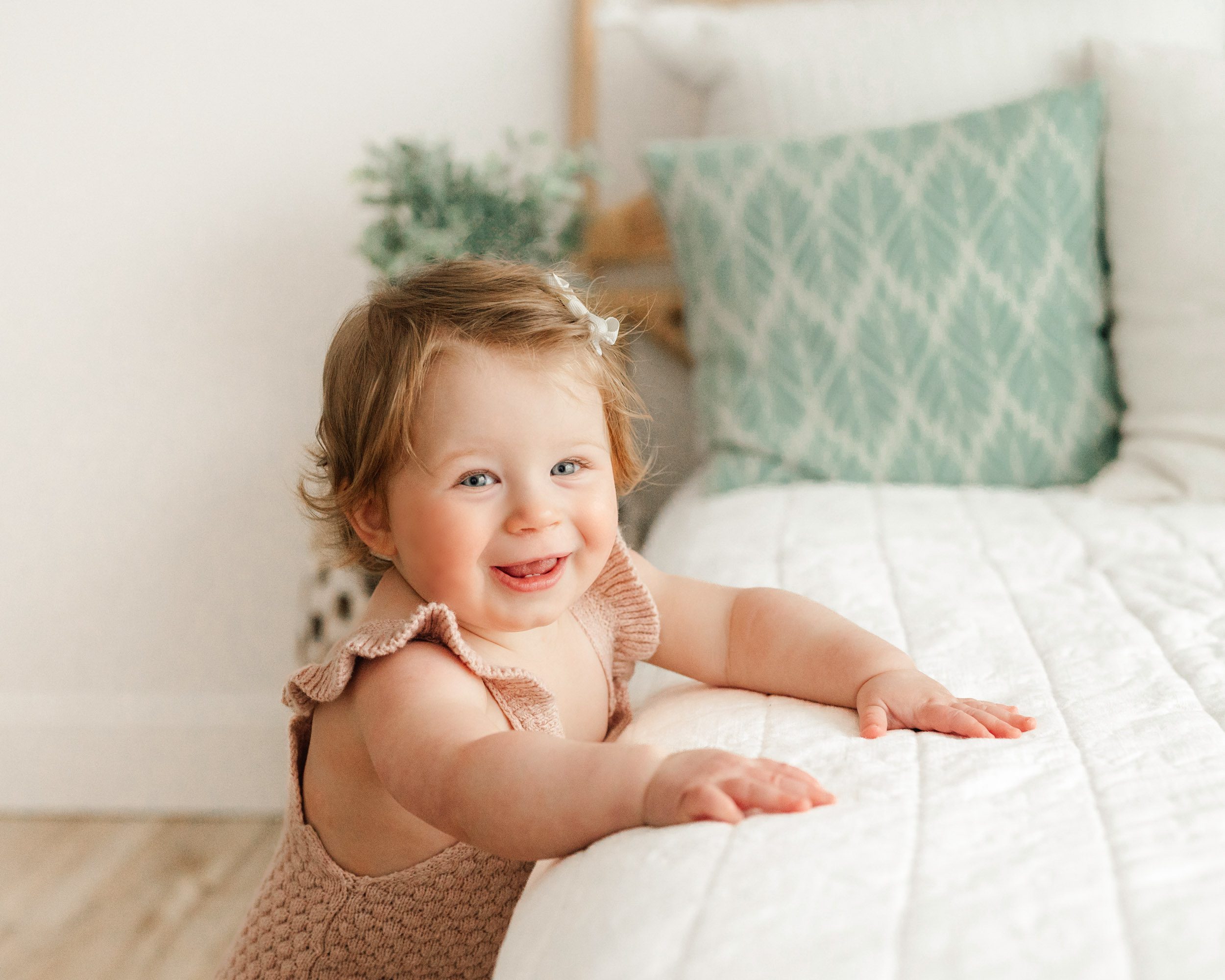 a baby girl standing and holding onto the edge of a bed as she looks at the camera with a huge smile on her face during a baby milestone photoshoot
