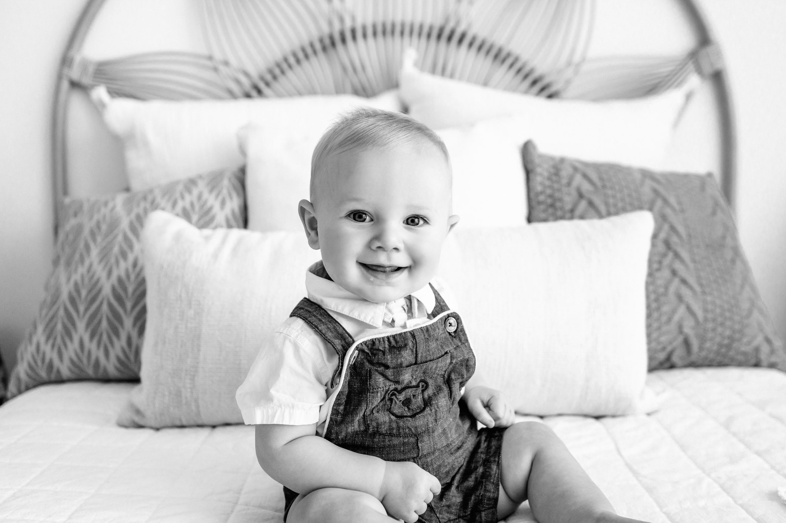 a black and white picture of a baby boy sitting on a bed and smiling at the camera during a Pottstown baby milestone photography session
