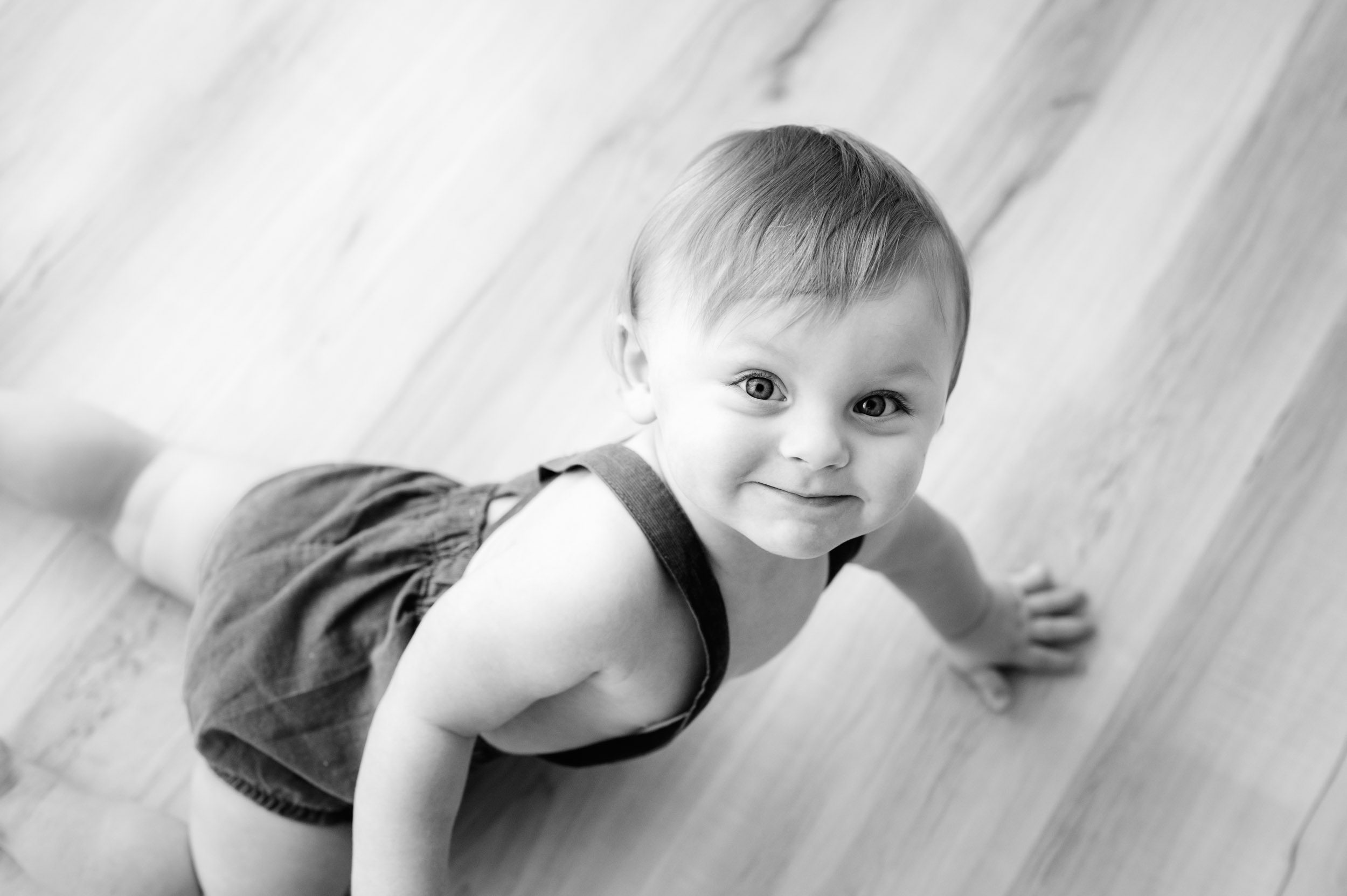 a black and white picture taken from above of a baby boy looking up at the camera and smiling as he crawls across the floor during a 1st birthday milestone photoshoot