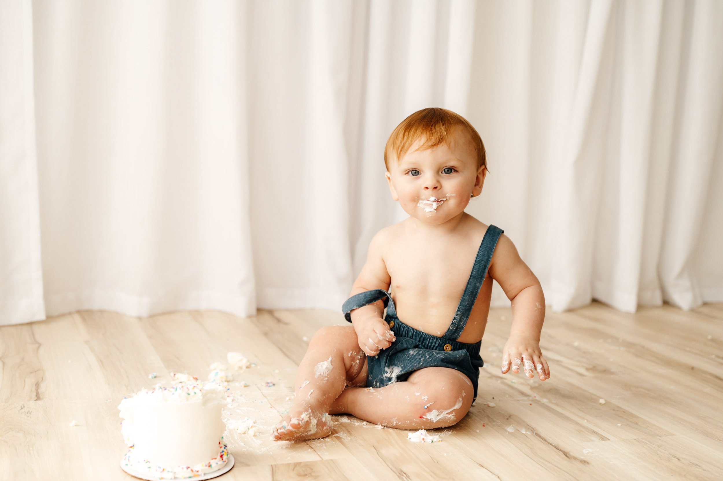 a boy sitting on the floor next to a small white cake with a big mess of cake all over his body as he smiles at the camera during a 1st birthday cake smash photoshoot
