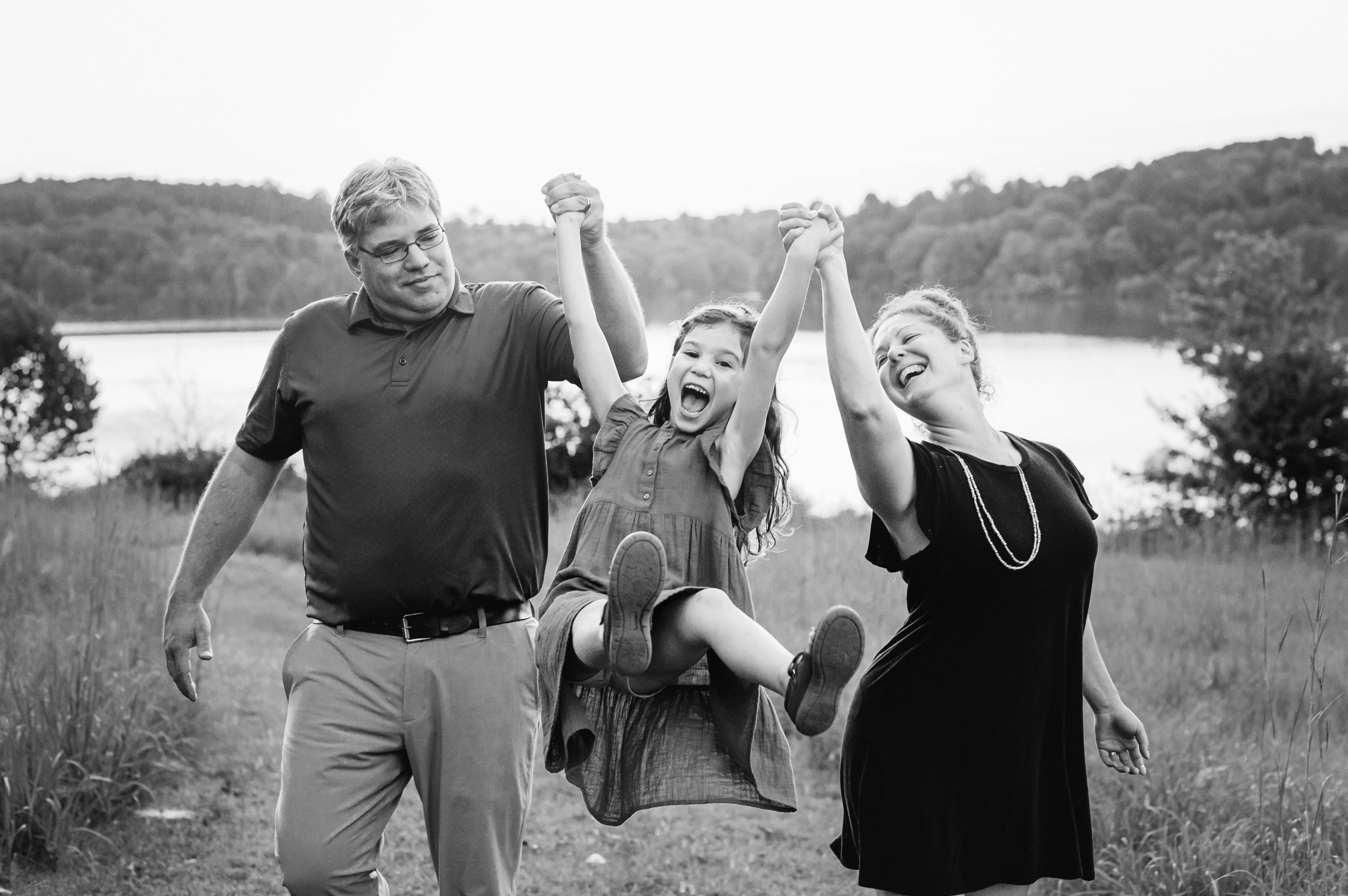a black and white picture of a young girl holding her parents' hands and swinging up in the air with a huge smile on her face during a family photoshoot