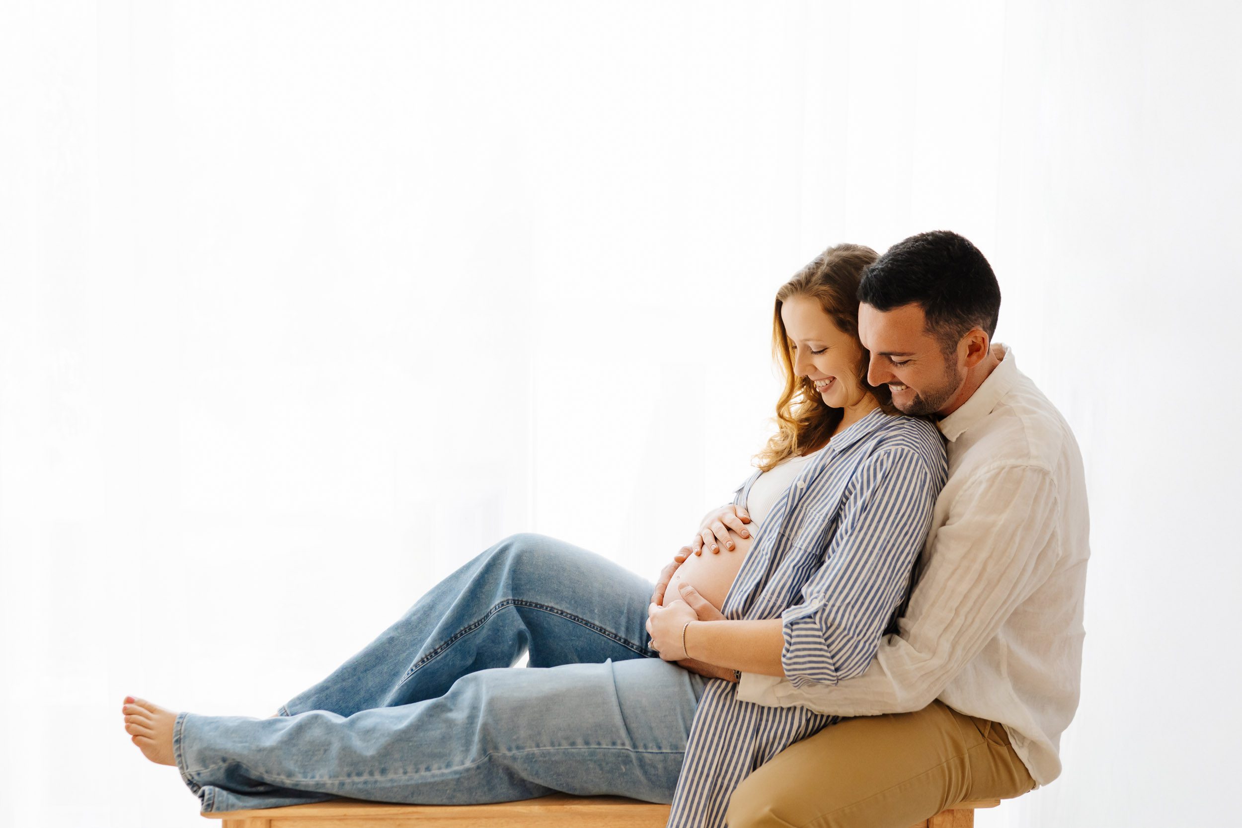 a backlit photo of expecting parents sitting on a bench and snuggling as they both smile down at mom's belly during a lifestyle maternity photography session