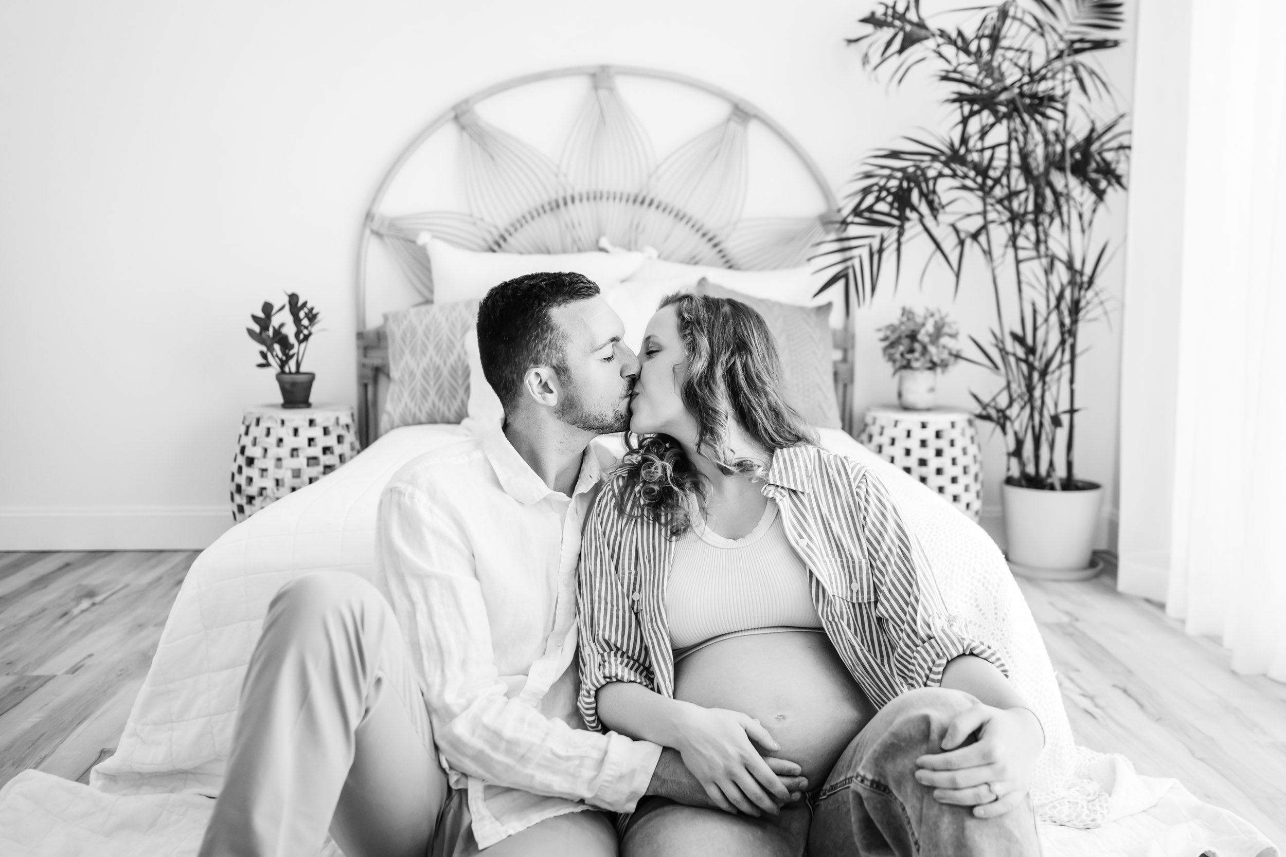 expecting parents sitting on the floor at the foot of a bed and giving each other a kiss as they both touch mom's belly during a lifestyle maternity photography session