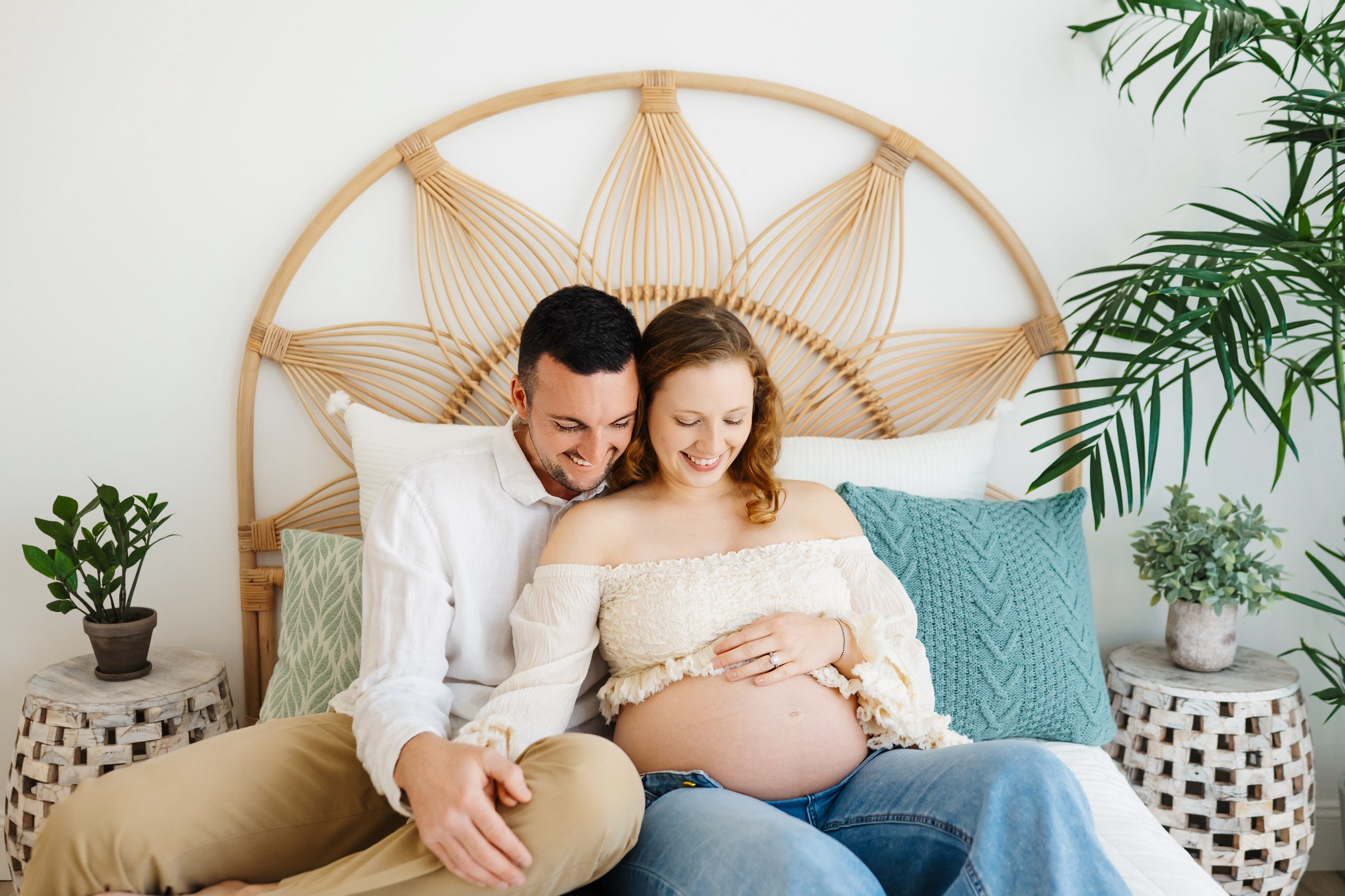 expecting parents sitting on a bed and snuggling together as they both smile down at mom's belly during a lifestyle maternity photography session