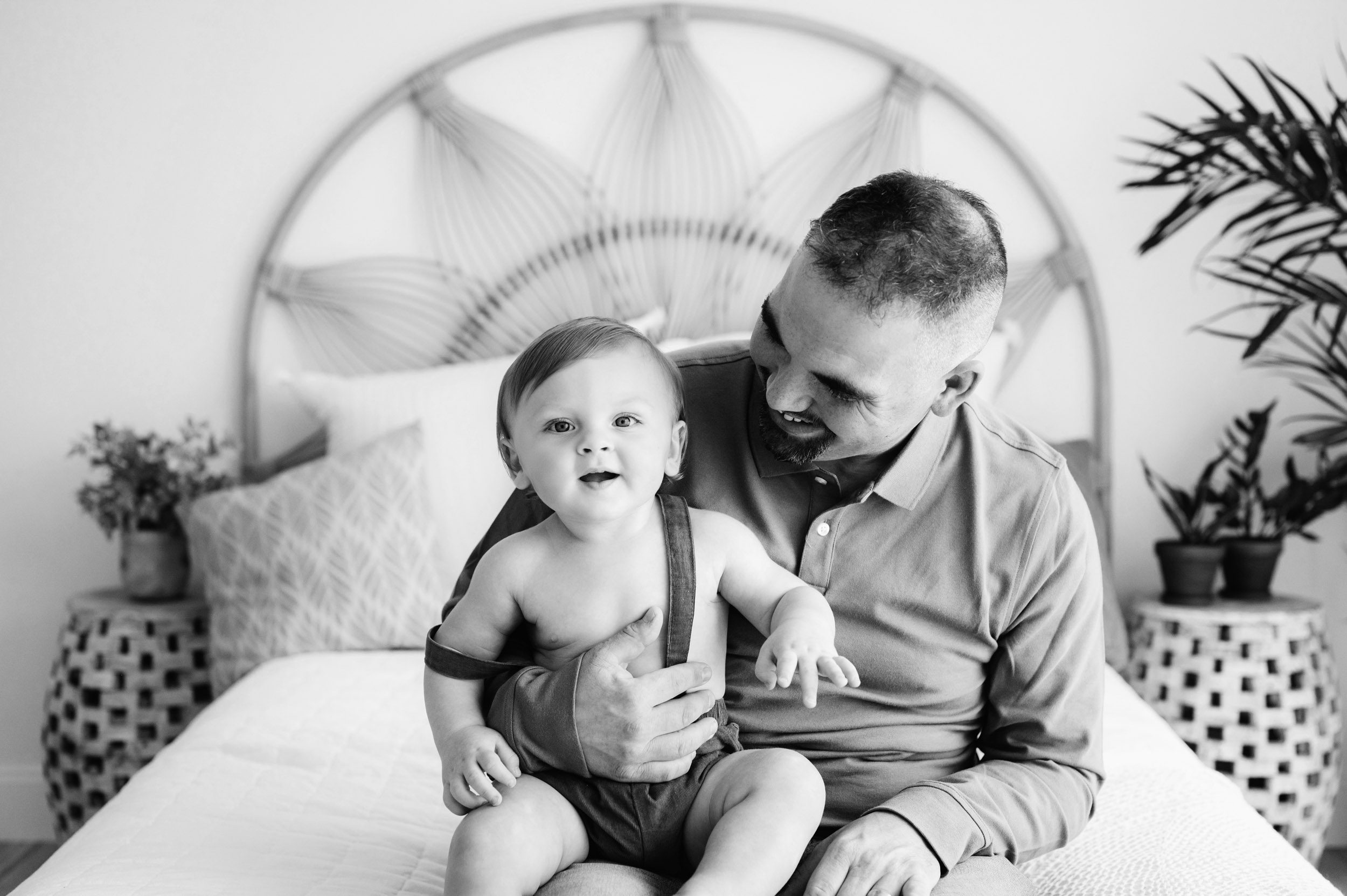 a black and white picture of a dad sitting on a bed with his young son and smiling at him during a 1st birthday photoshoot