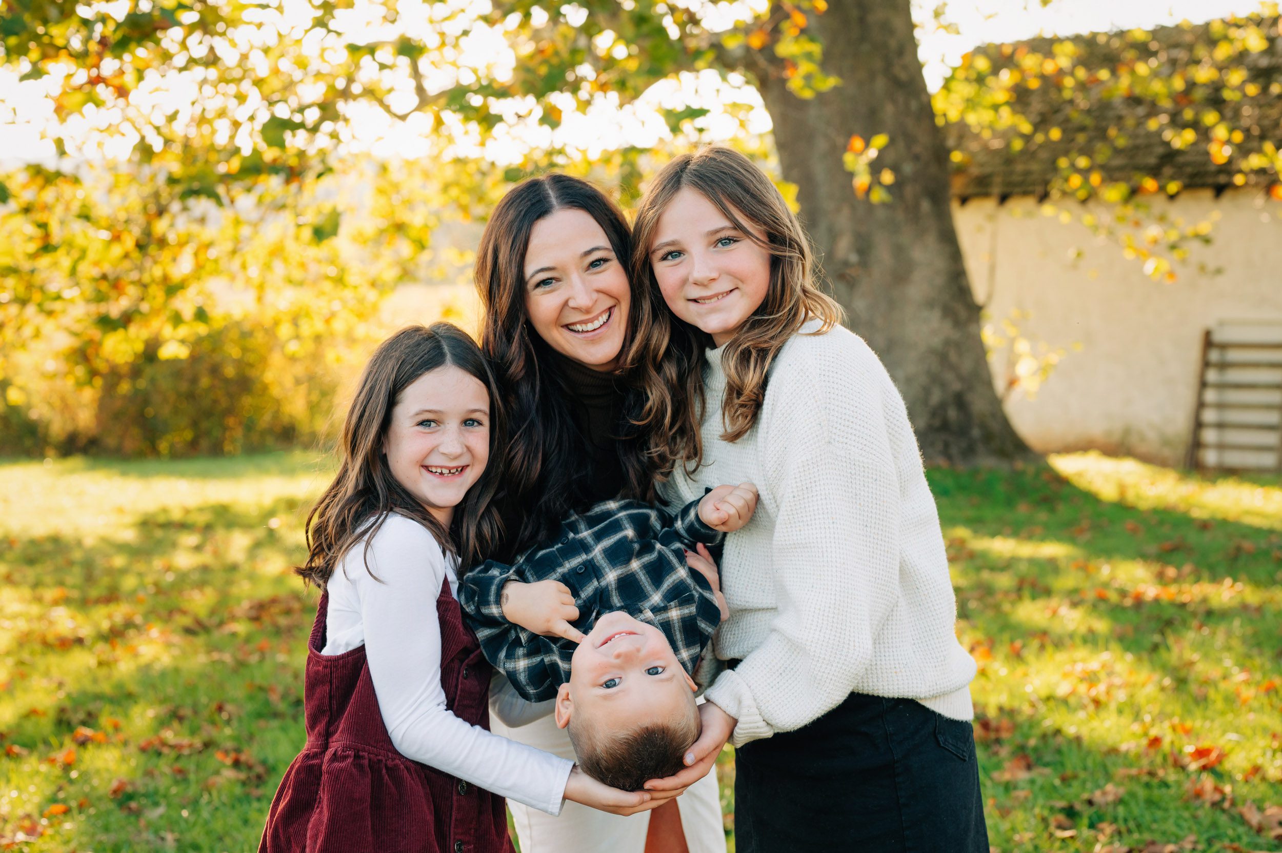 a mom holding her youngest son upside down while her two older daughters hug her from either side and they all smile at the camera during a family photoshoot