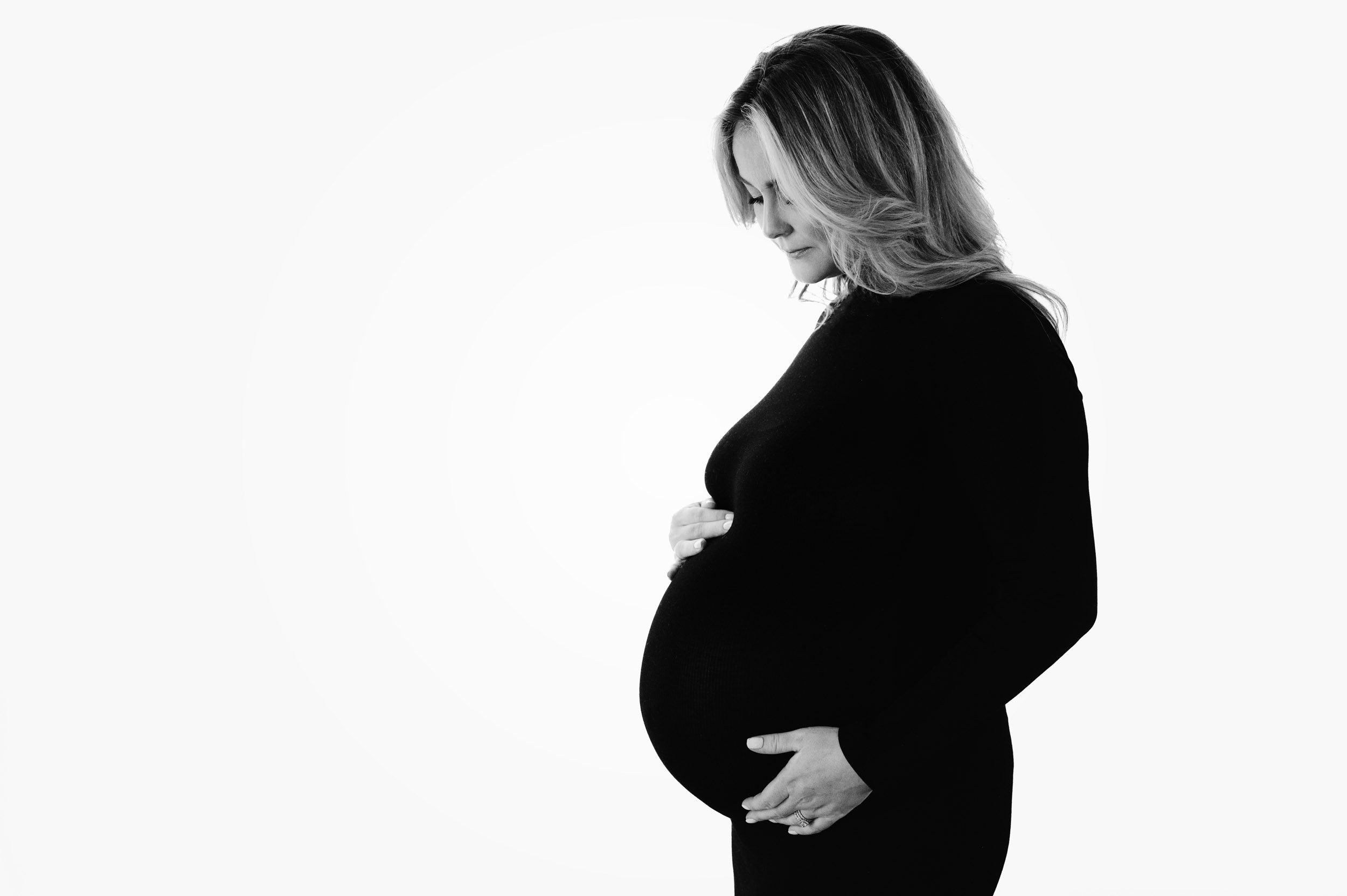 a black and white backlit photo of an expecting mom wearing a fitted black maternity dress and gently cradling her belly while she looks down at her belly 