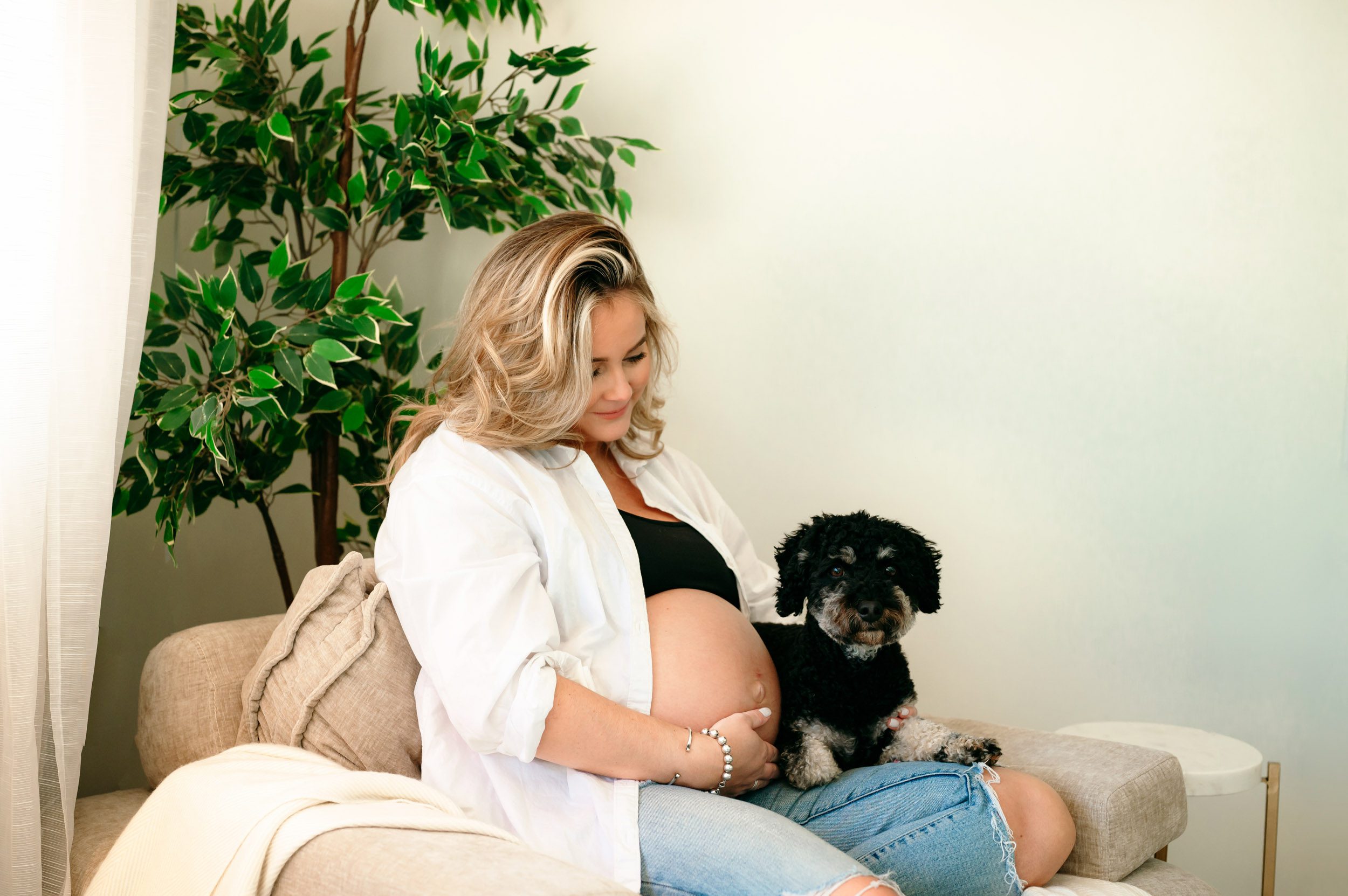 an expecting mom sitting on her belly holding her puppy on her lap and smiling down at him during an in home lifestyle maternity photoshoot