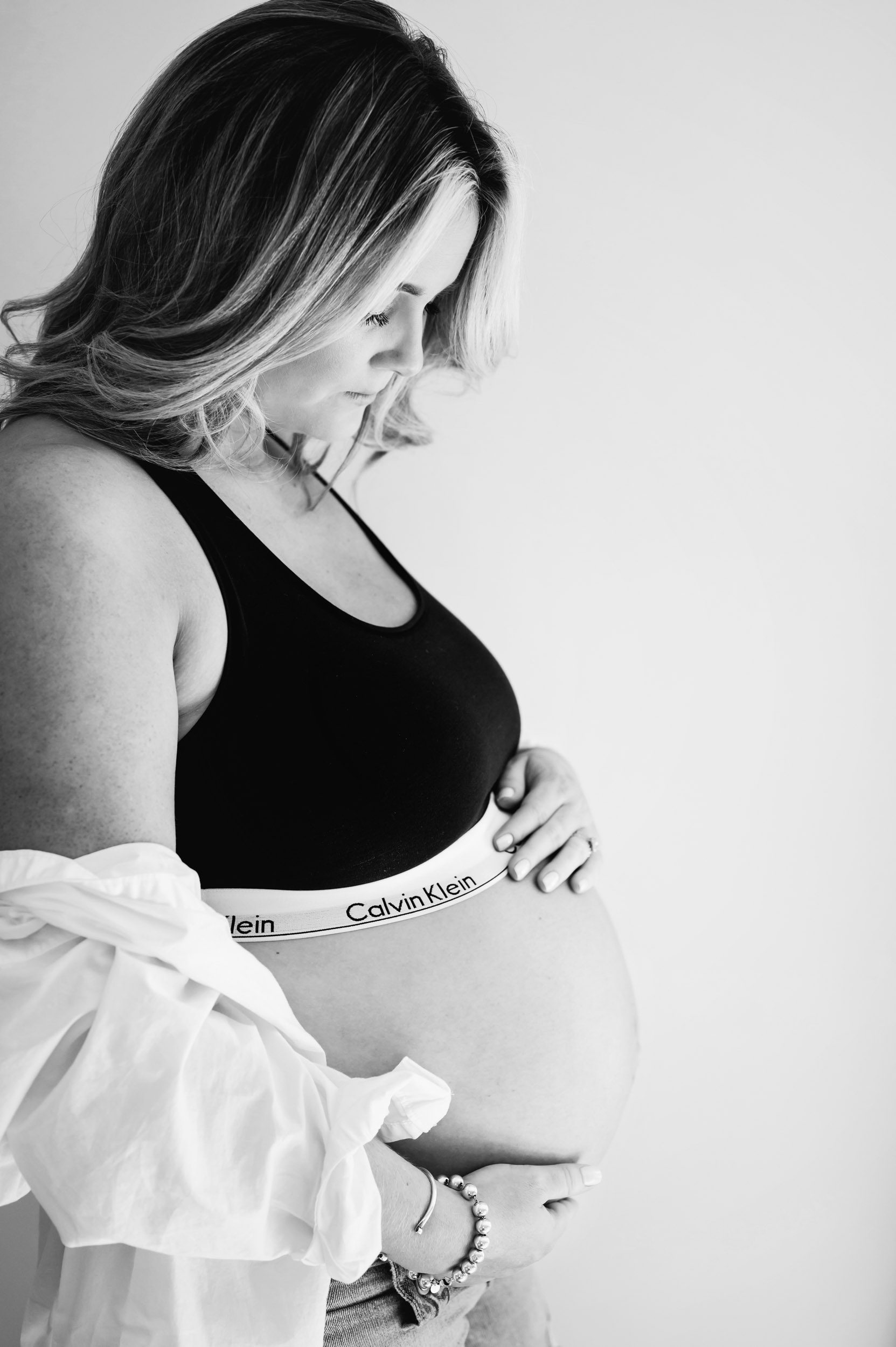 a black and white picture of an expecting mom wearing a black Calvin Klein bra and jeans as smiles down at her belly 