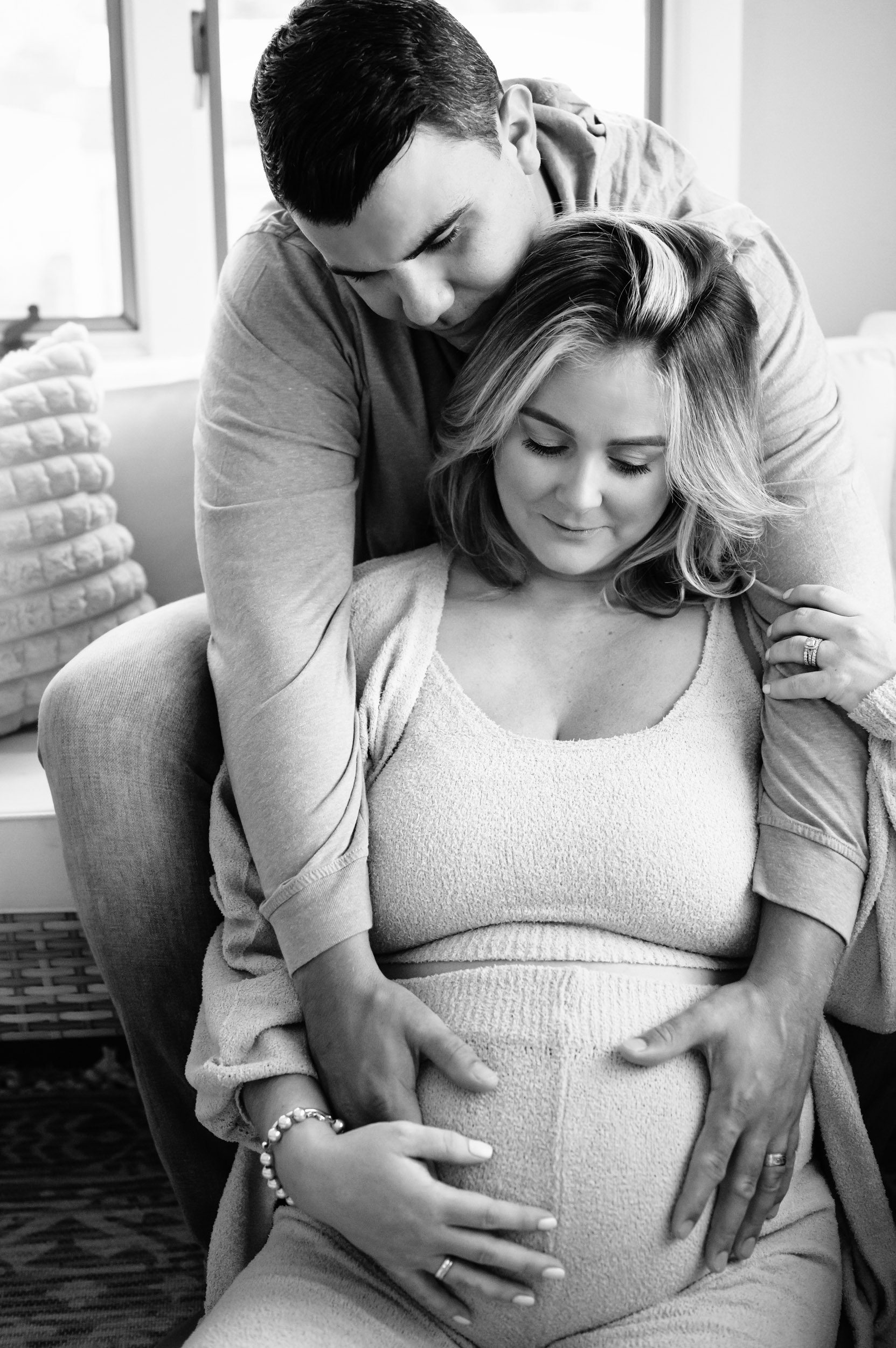 a black and white picture of an expecting mother sitting on the floor in front of the sofa while dad sits on the sofa behind her and leans over her shoulder to hug her and touch her belly during an in home lifestyle maternity photography session