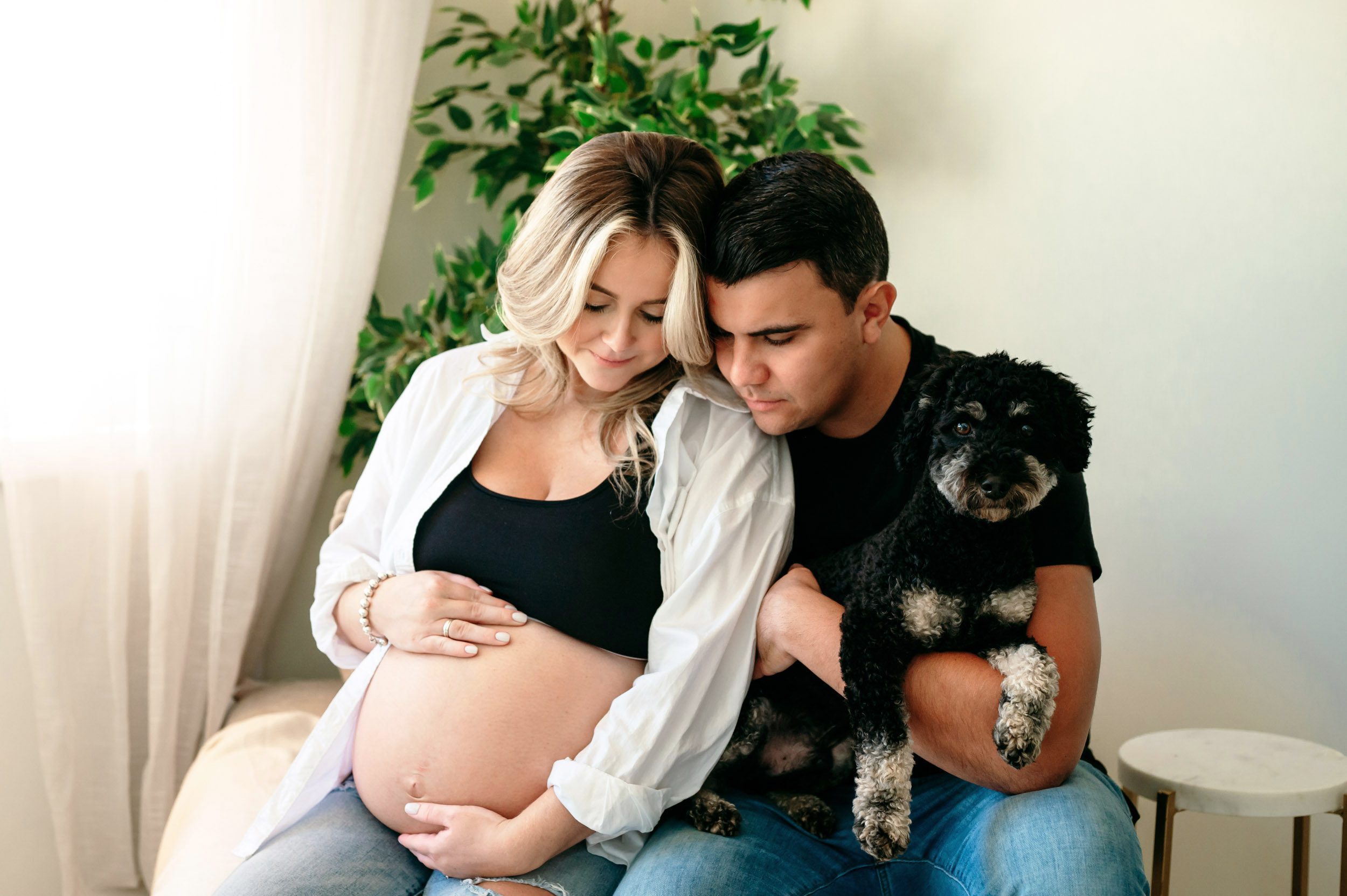 expecting parents sitting on an oversized chair as they both look down at mom's belly while their pet dog sits in dad's lap and looks directly at the camera during an in home lifestyle maternity photoshoot