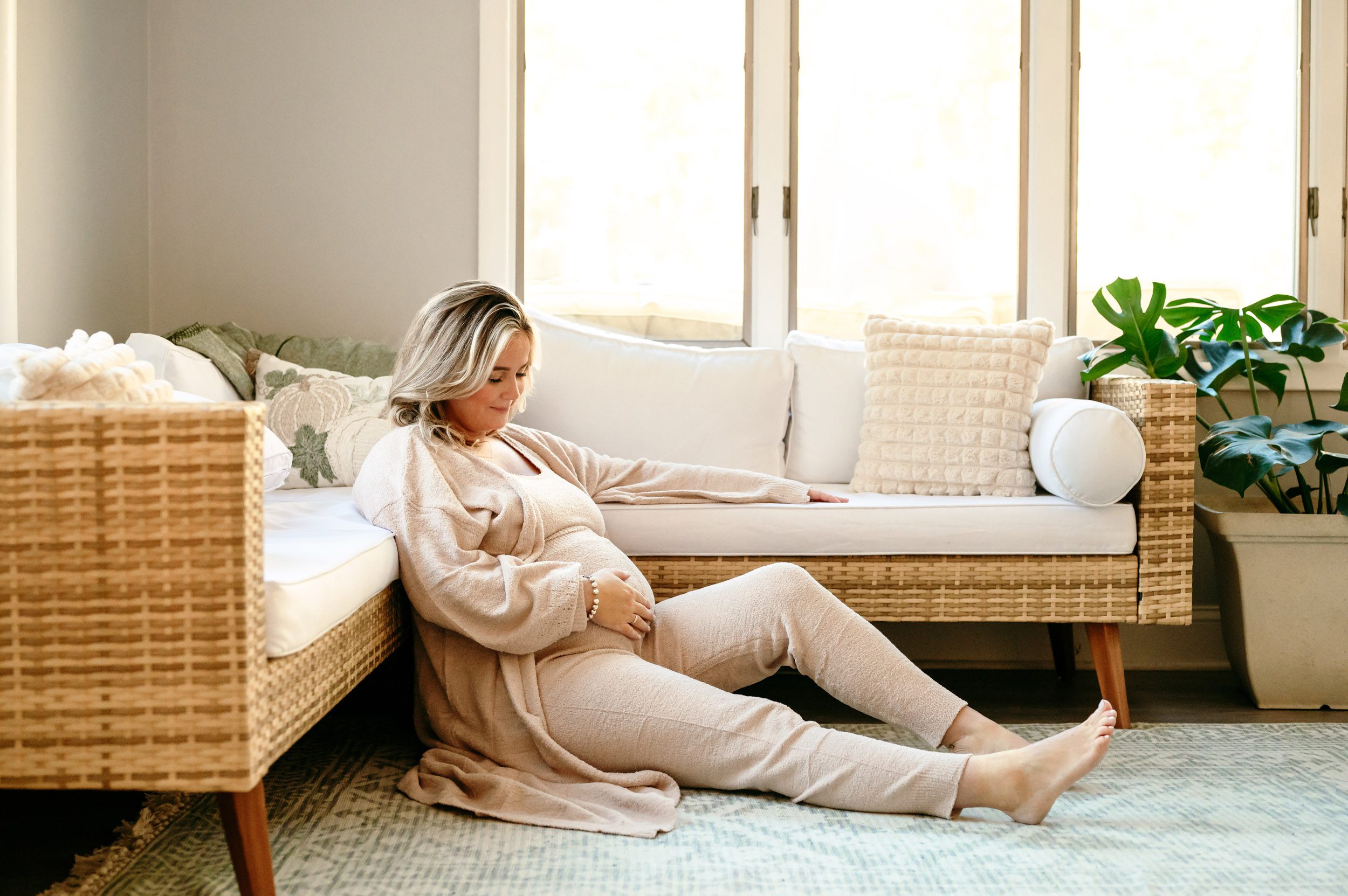 an expecting mom sitting on the floor in front of a wicker sofa in her sunroom smiling down at her belly during a lifestyle maternity photoshoot