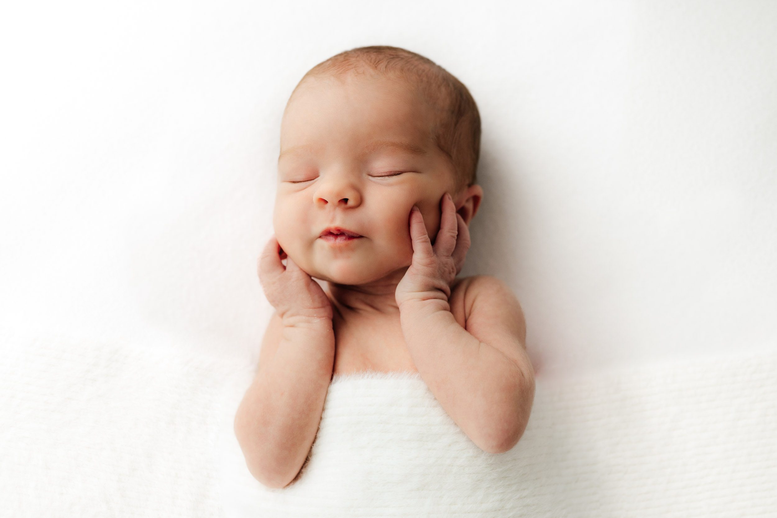 a baby girl laying on a white backdrop under a fuzzy white blanket holding her hands up to both of her cheeks and pursing her lips during a newborn photoshoot