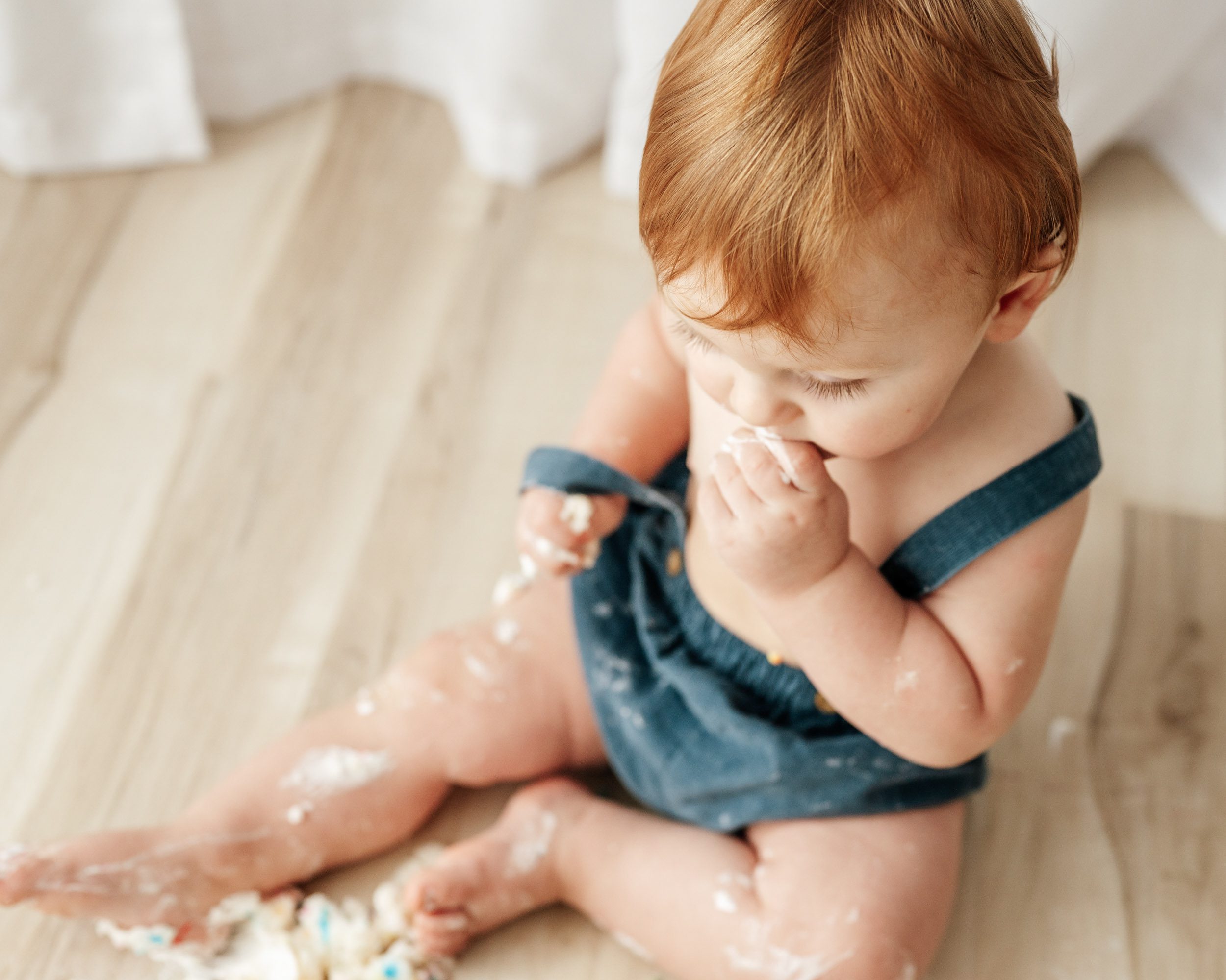a picture taken from above of a young boy wearing blue shorts with suspenders putting a bite of cake in his mouth with his hand during a 1st birthday cake smash photoshoot