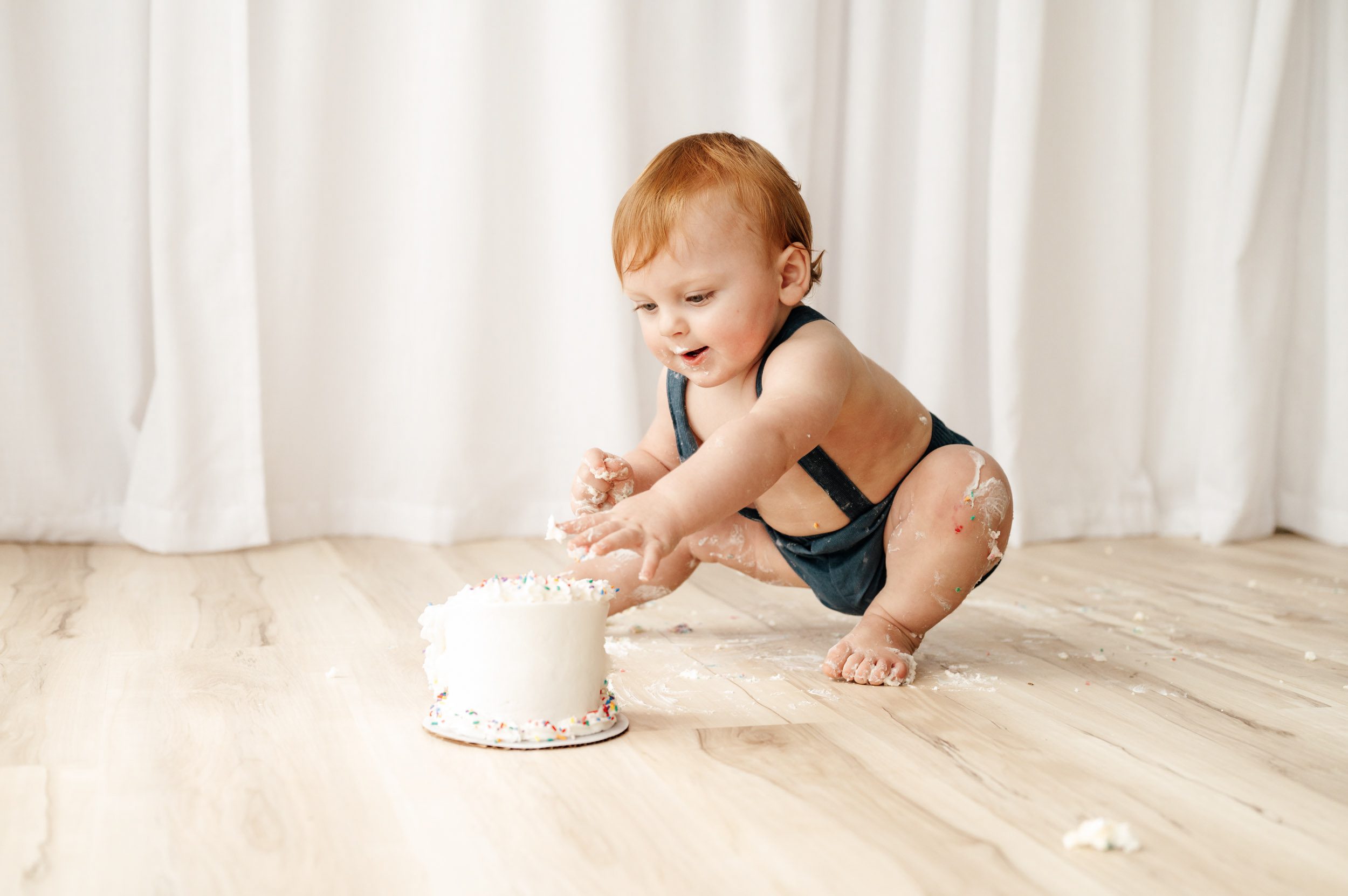 a young boy wearing blue shorts with suspenders squatting down in front of a small white cake and reaching out to touch the icing with his hand during a 1st birthday cake smash photoshoot
