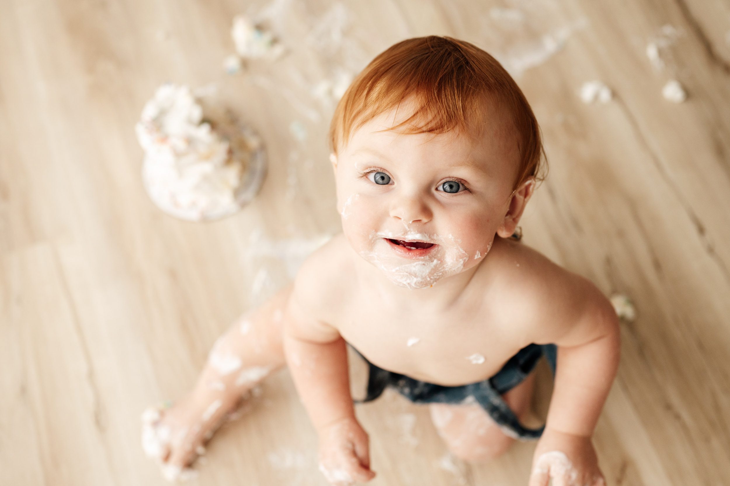 a picture taken from above of a boy with cake all over his face looking straight up at the camera and smiling during a 1st birthday cake smash photoshoot