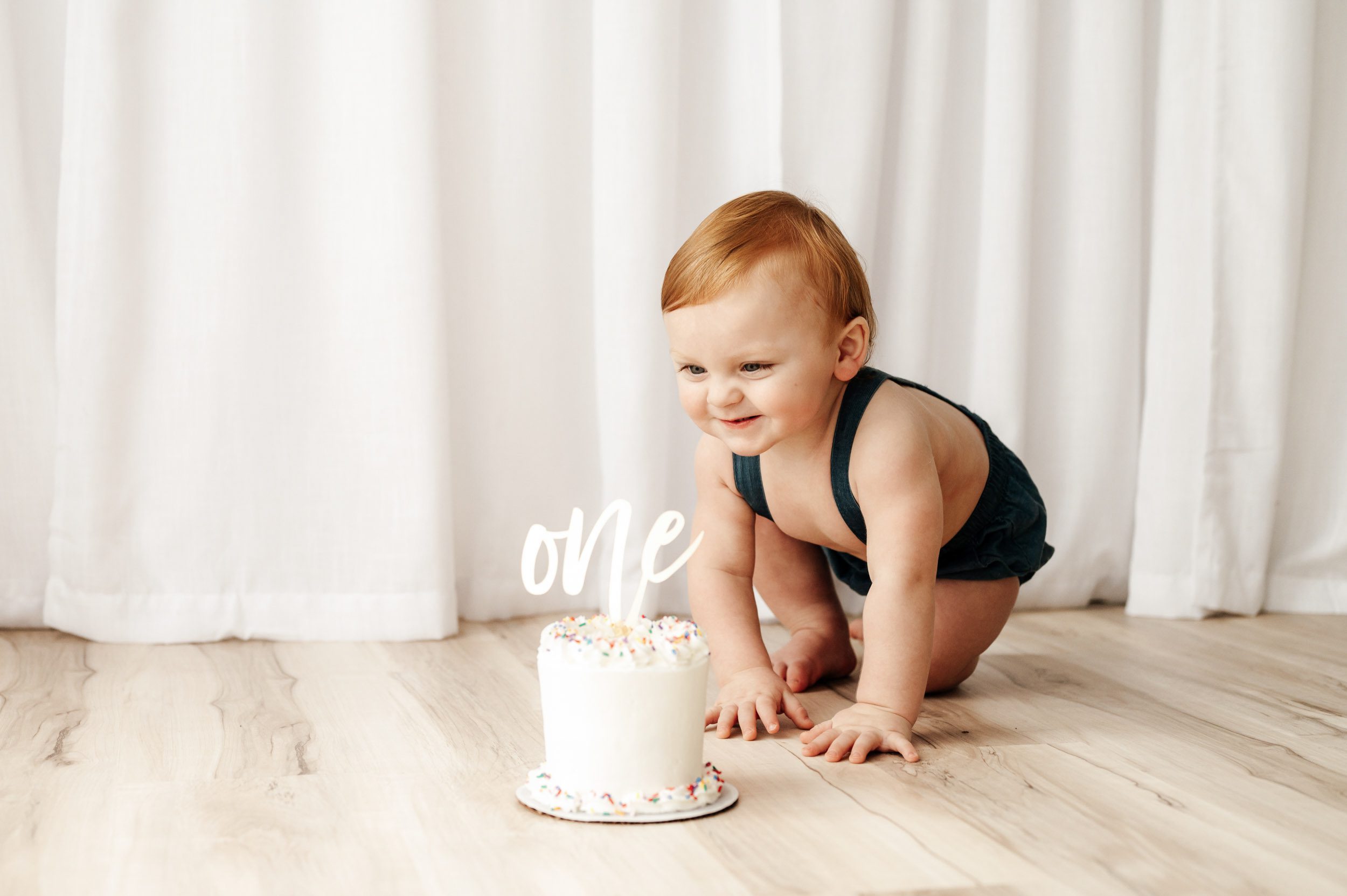 a young boy crawling across the floor toward a small white cake with the letters ONE on top and scrunching up his nose with excitement as he gets ready to reach out and touch the cake during a 1st birthday cake smash photoshoot