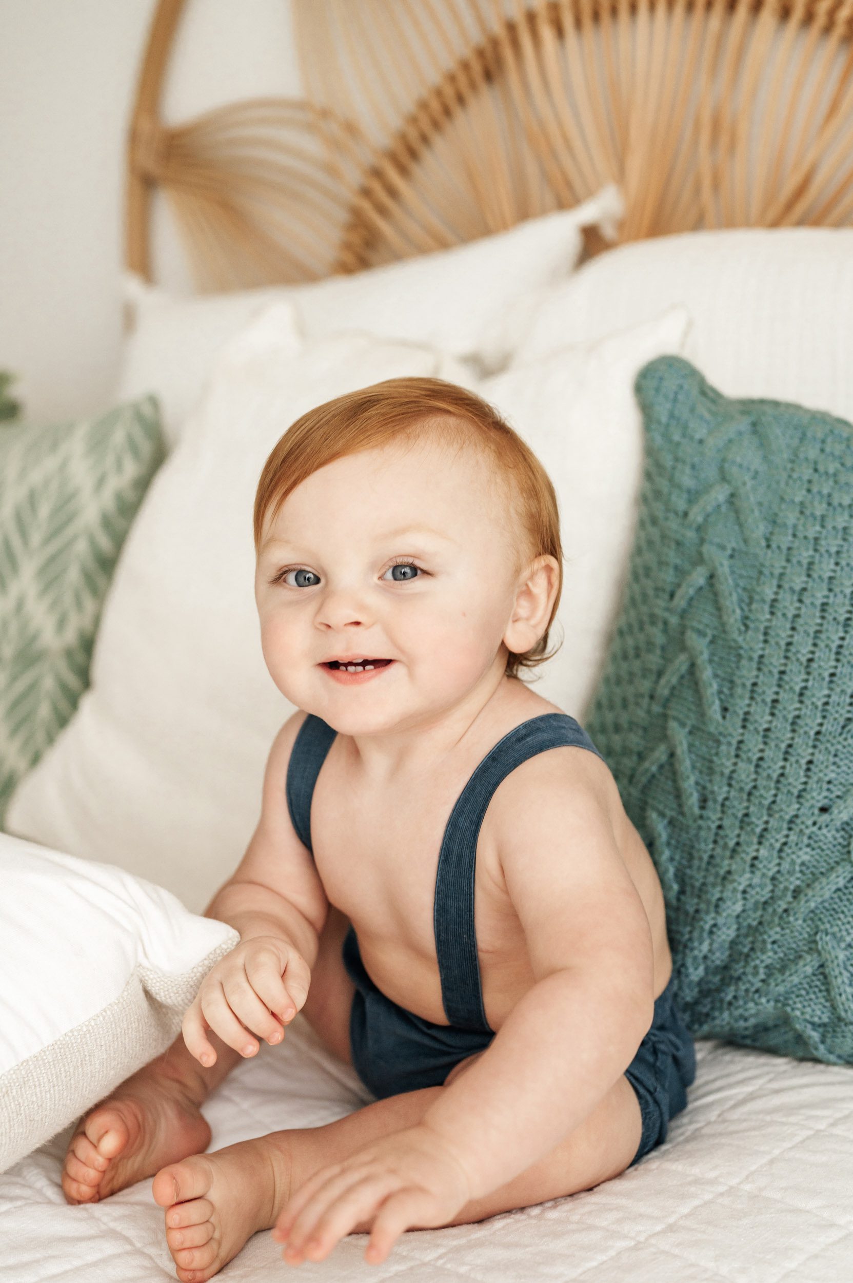 a young boy wearing blue shorts with suspenders sitting on a bed and smiling at the camera during a milestone photo session