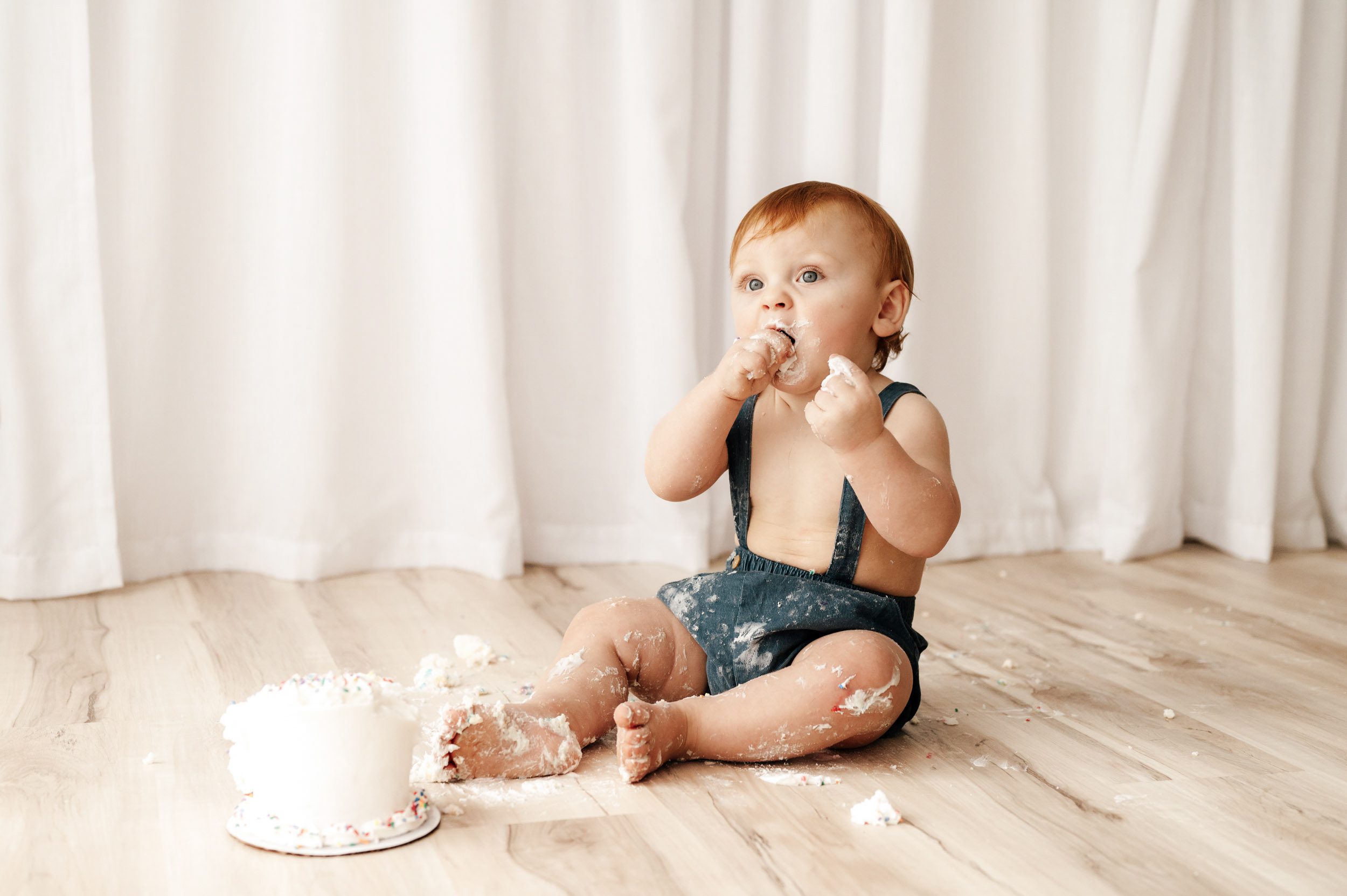 a young boy wearing blue shorts with suspenders sitting on the floor in front of a small white cake with cake all over his arms and legs and the floor in front of him as he puts a bite of cake into his mouth with his hand during a 1st birthday cake smash photoshoot