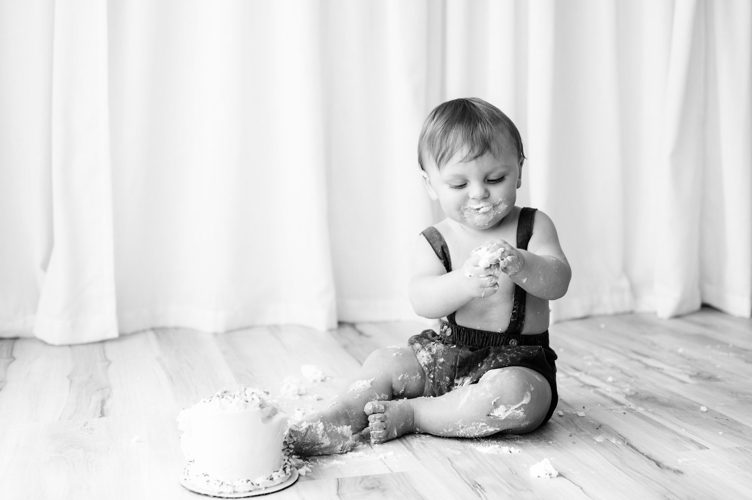 a black and white picture of a young boy sitting on the floor with a small white cake in front of him as he smashes the cake with his fingers during a 1st birthday cake smash photoshoot