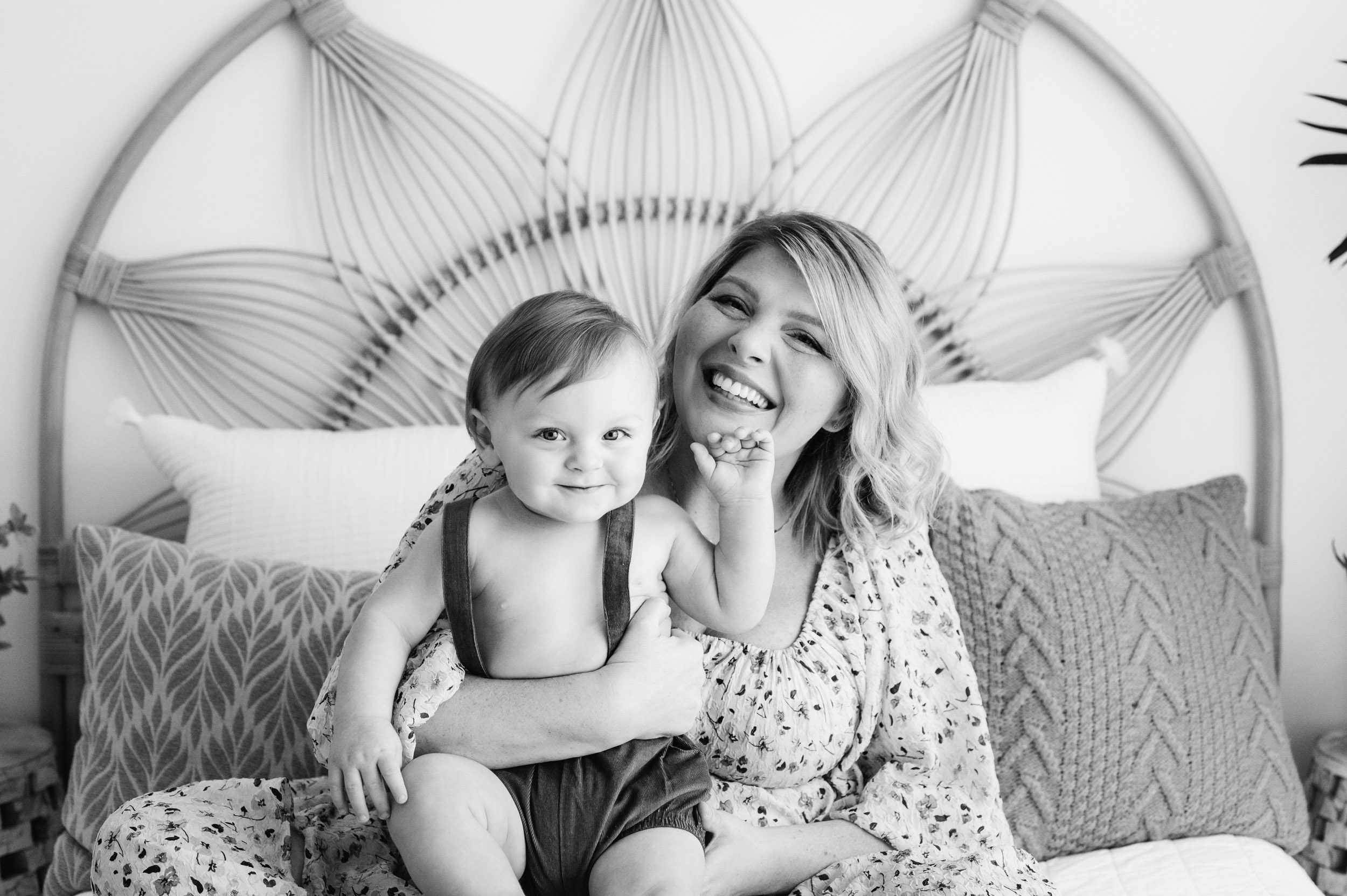 a black and white picture of a mom sitting on a bed snuggling her young son on her lap as they both look at the camera and smile during a 1st birthday photoshoot