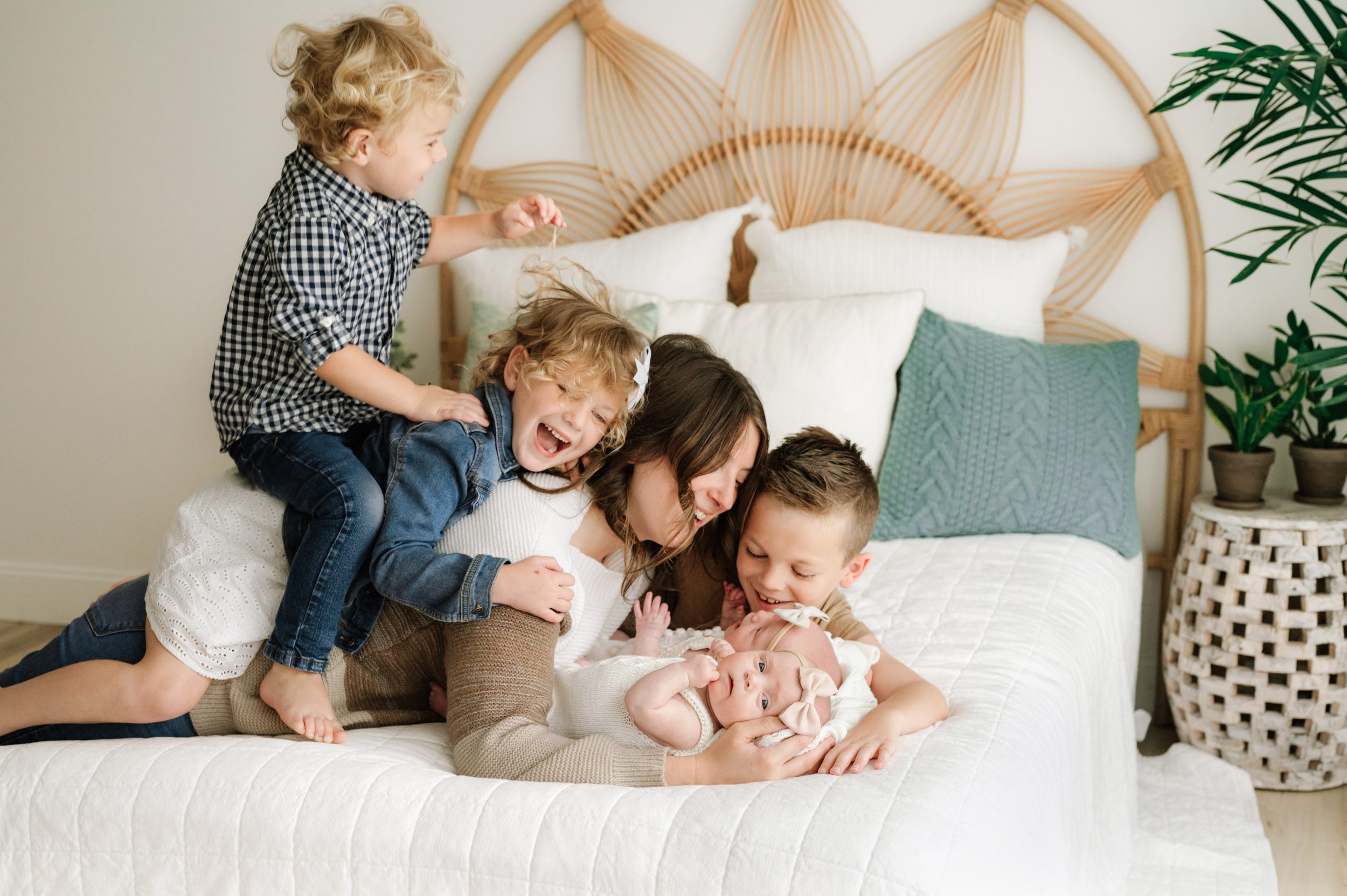 a mom laying on the bed with her twin baby girls and her three older children snuggled around her and jumping on her back as they all laugh during a lifestyle newborn photoshoot