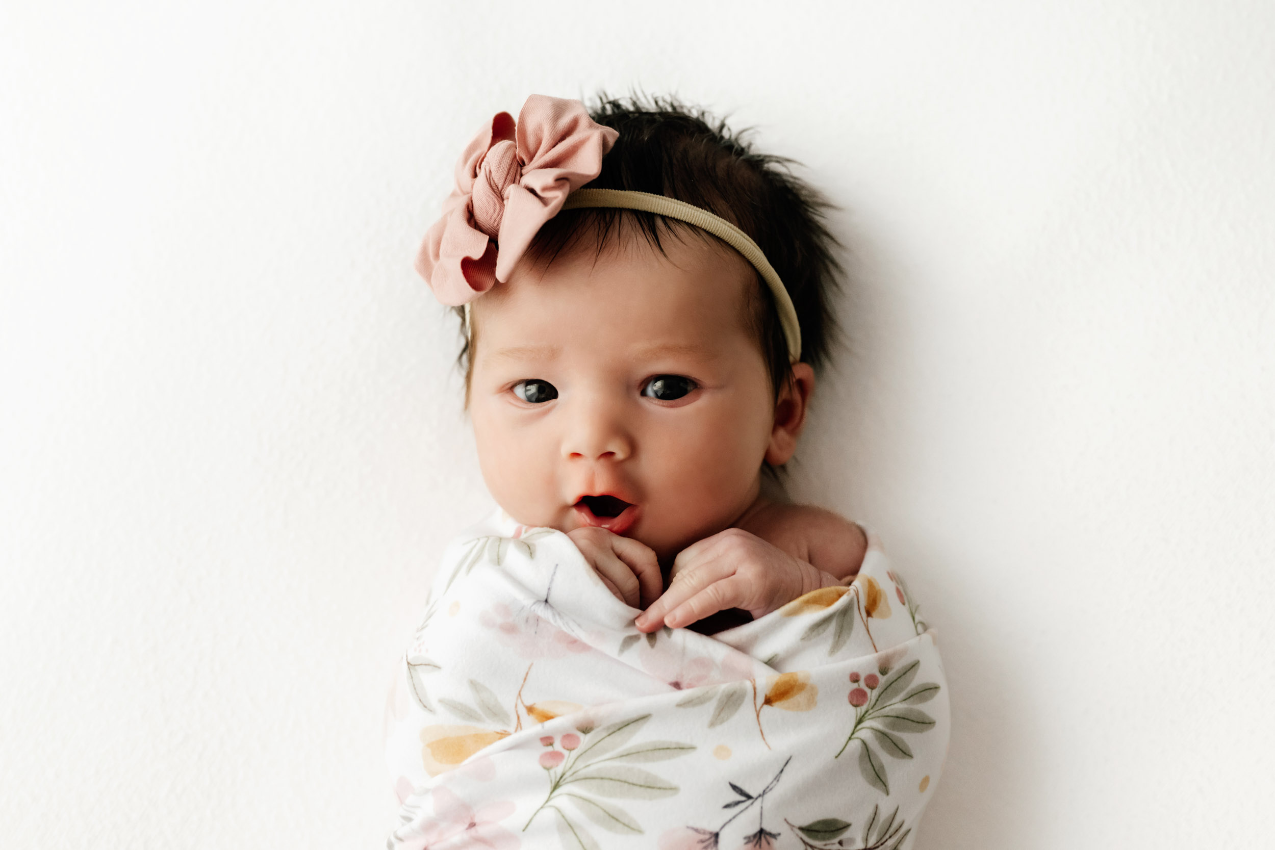 a baby girl wrapped in a floral swaddle blanket wearing a pink headband and looking directly at the camera during a natural newborn photos session