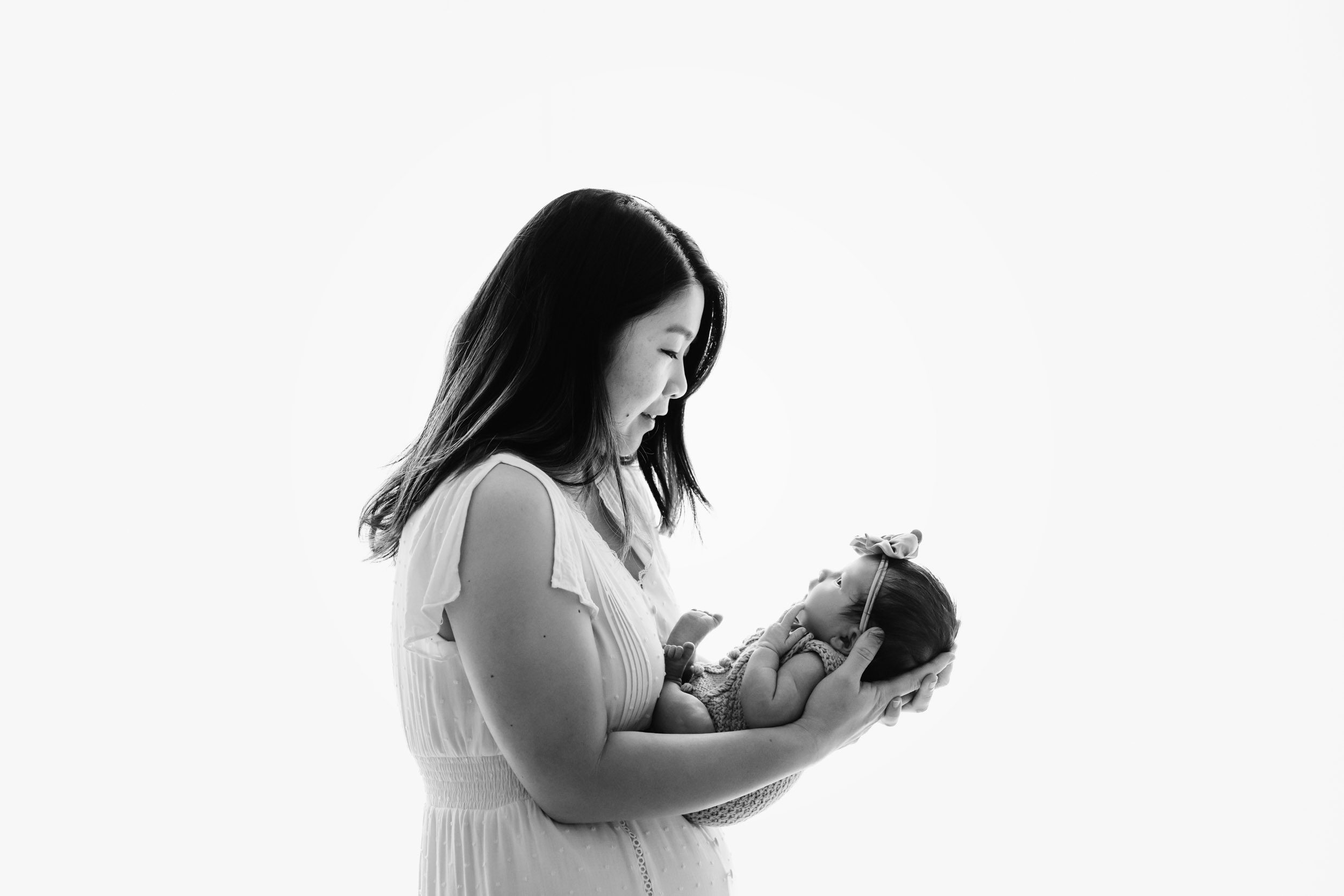 a black and white backlit photo of a new mom holding her baby girl in her arms and smiling down at her during a natural newborn photoshoot