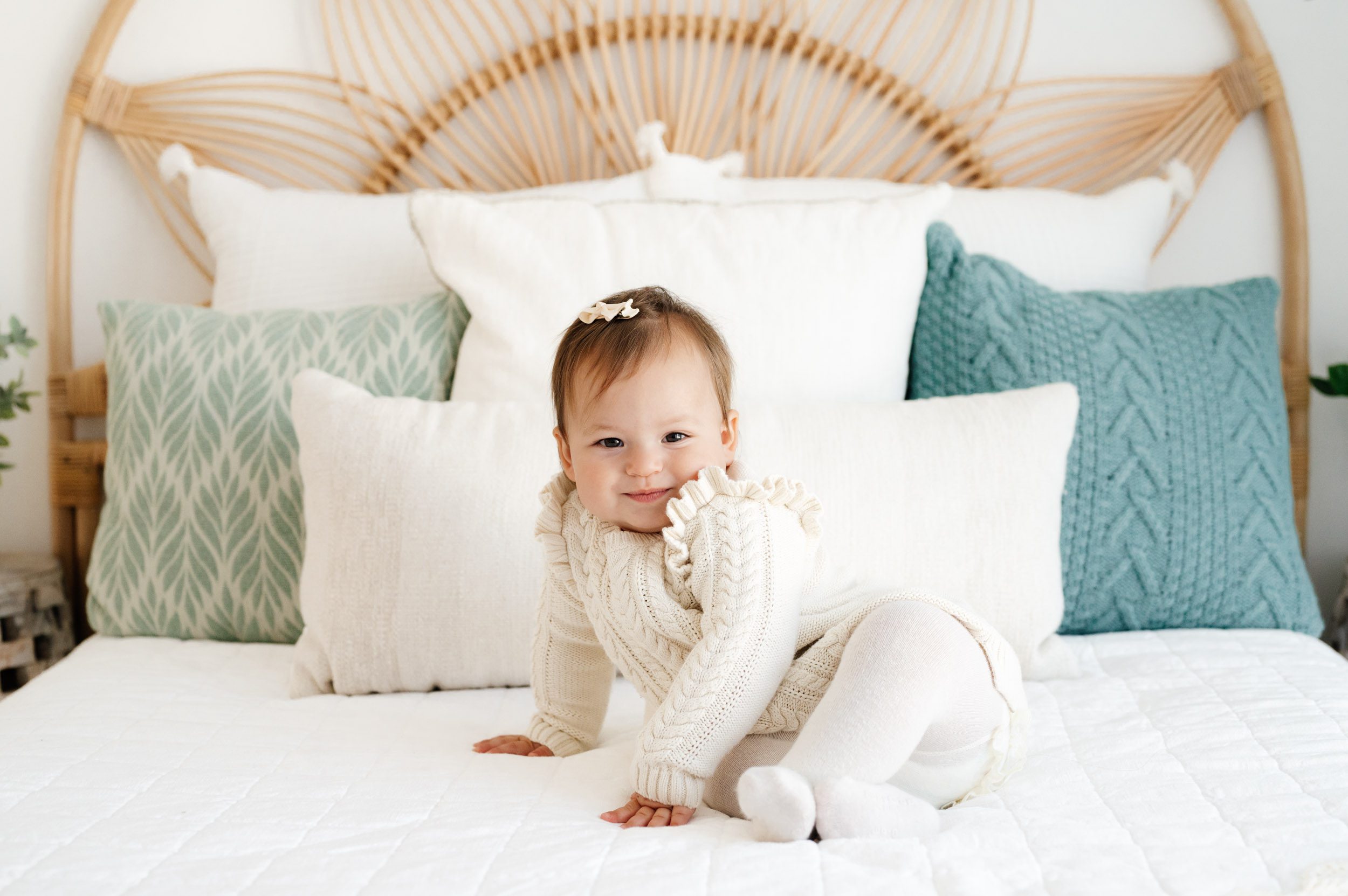 a little girl wearing an ivory cable knit jumper and tights sitting on a bed and looking right at the camera with a smirky smile on her face during a Pottstown baby photography session