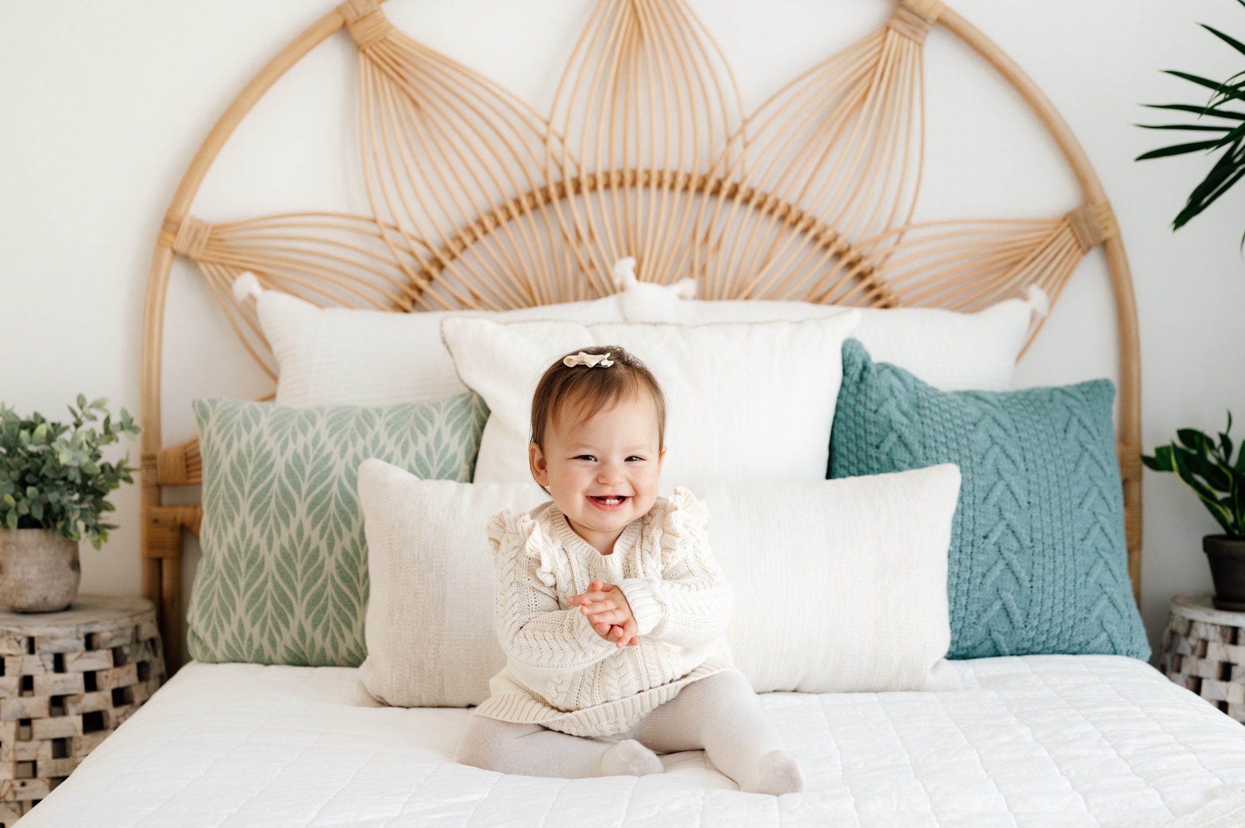 a little girl wearing an ivory cable knit jumper and tights sitting on a bed and smiling right at the camera as she claps her hands during a Pottstown baby photography session