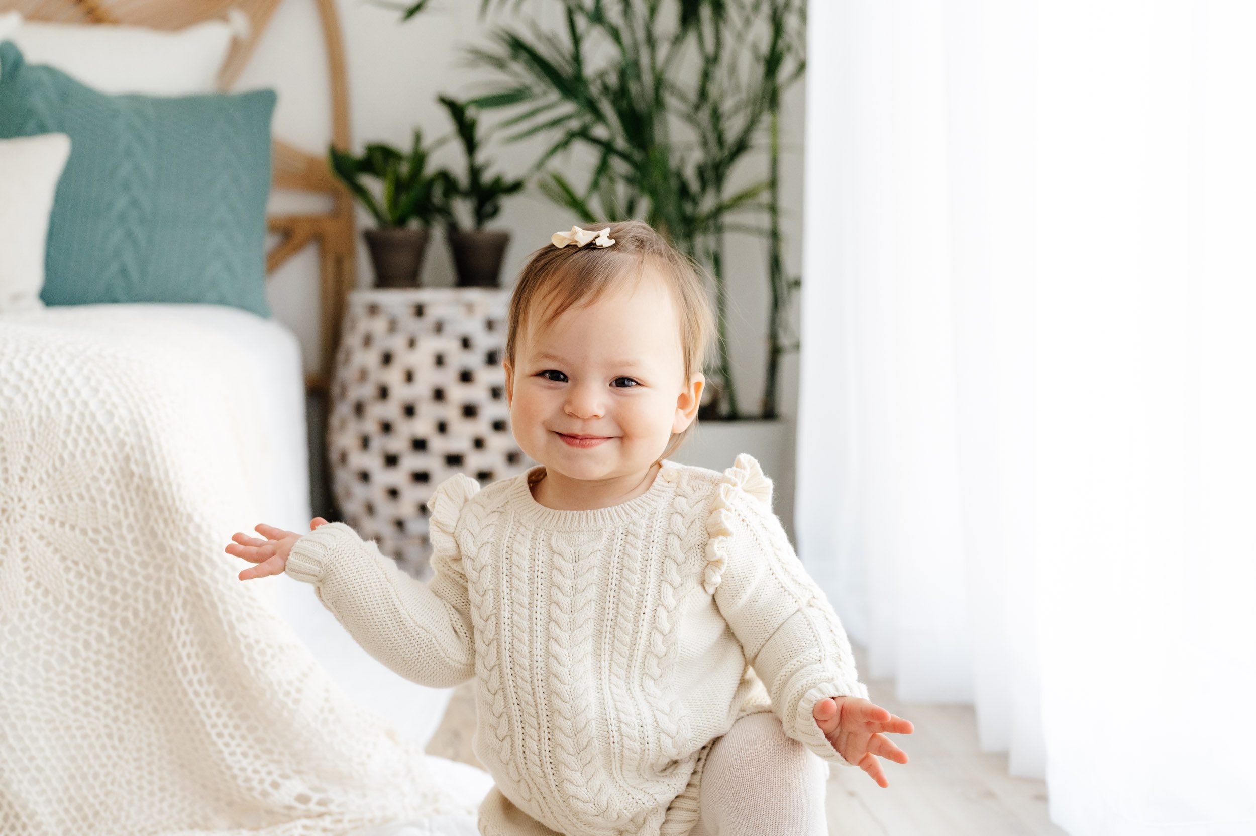 a little girl wearing an ivory cable knit jumper kneeling down next to a bed and smiling at the camera as she holds her arms out to the sides during a Pottstown baby photography session