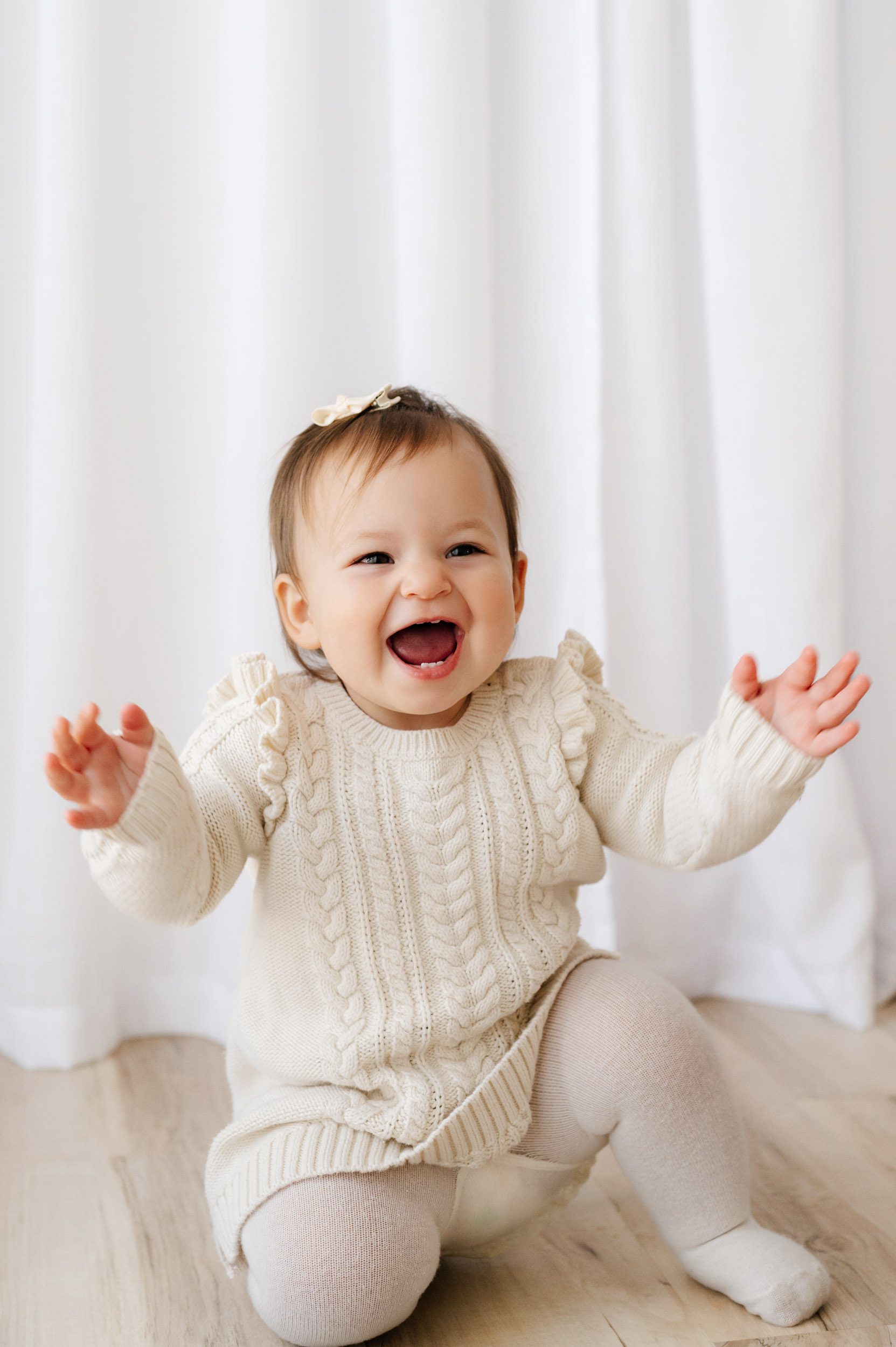 a little girl wearing an ivory cable knit jumper  and tights squatting down on the floor and looking up at her mom with a huge smile on her face during a Pottstown baby photography session