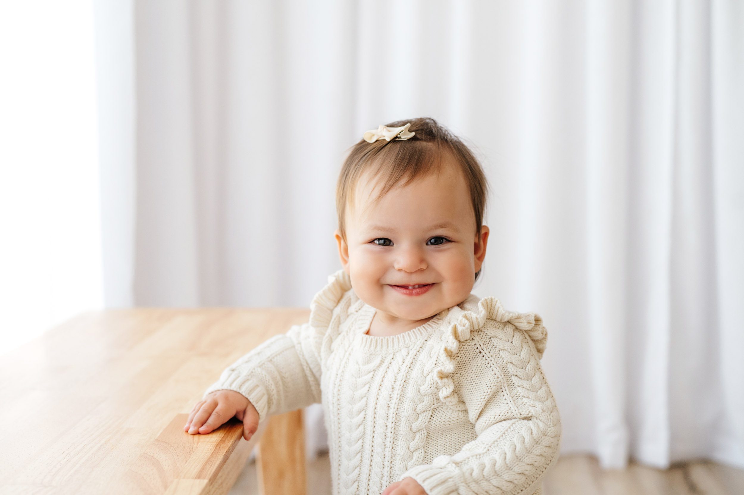 a little girl wearing an ivory cable knit jumper dress standing and holding onto a wood bench as she looks directly at the camera with a toothy grin during a Pottstown baby photography session