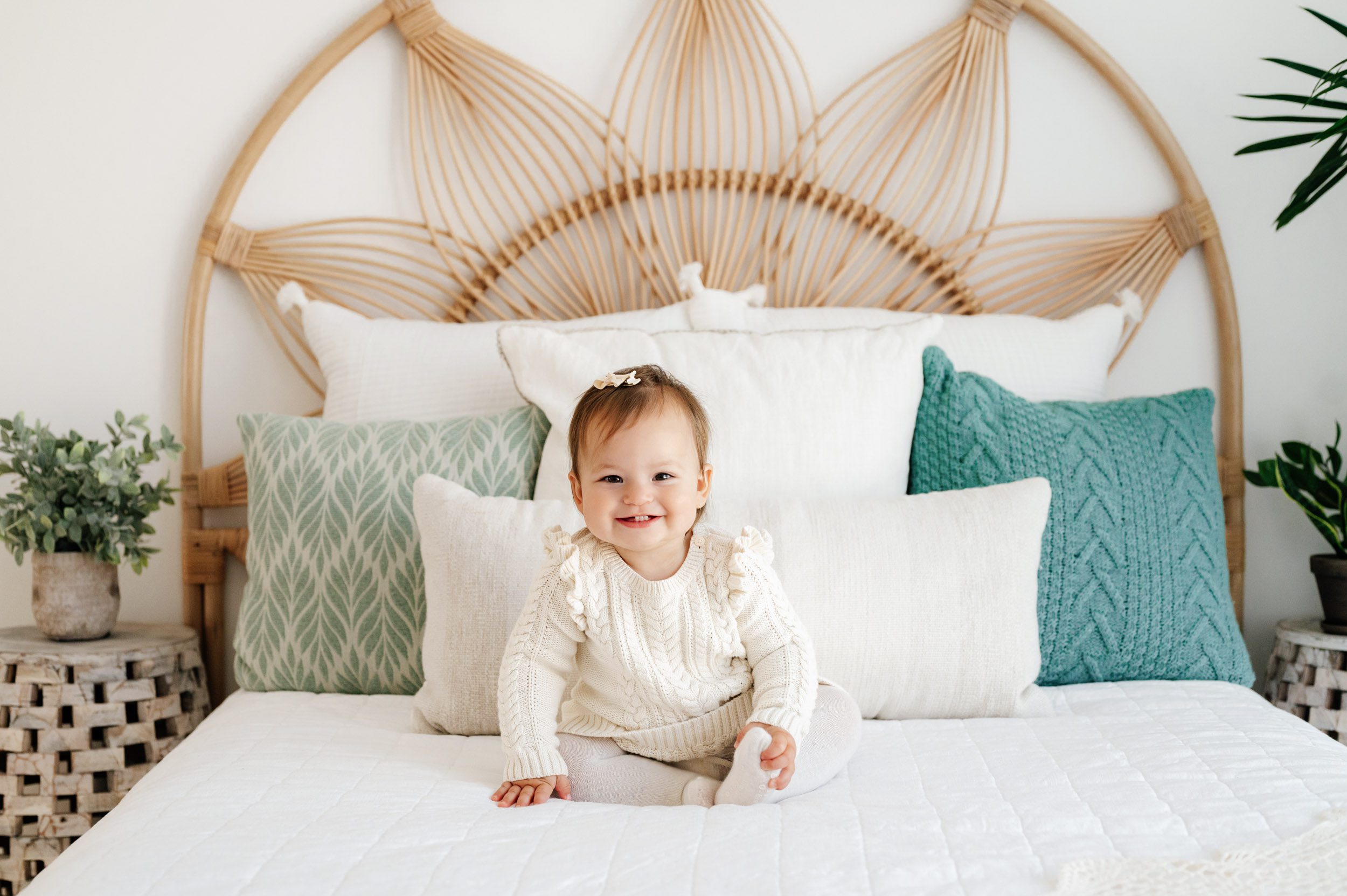 a little girl wearing an ivory cable knit jumper and tights sitting on a bed and smiling right at the camera as she grabs her foot with her hand during a Pottstown baby photography session