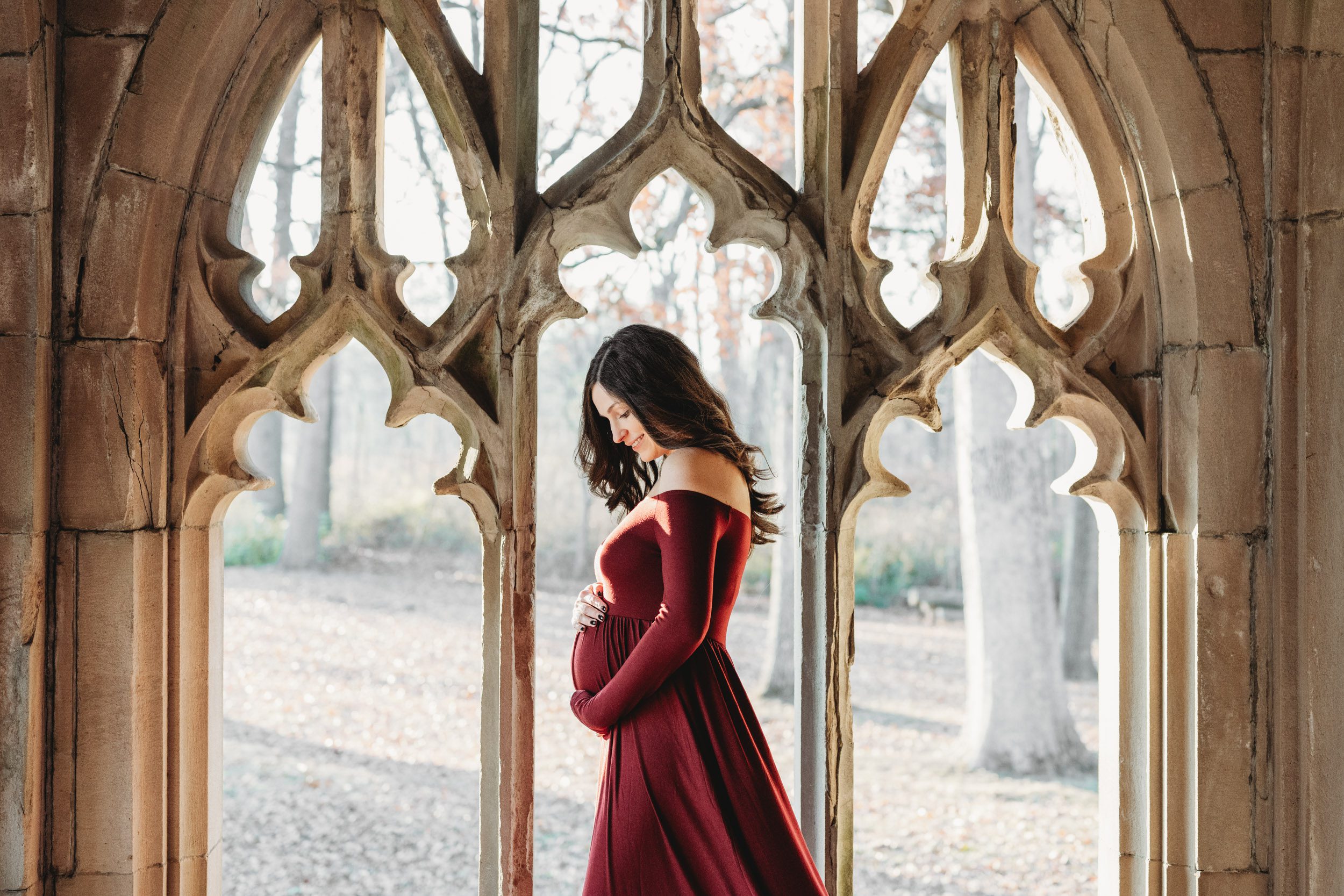 an expecting mom in a long sleeve wine colored dress standing in the middle of a stone archway and smiling down at her belly during a winter maternity photoshoot