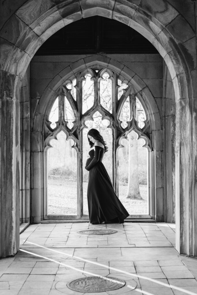 a black and white picture of an expecting mom standing in the middle of a stone archway and cradling her belly during a winter maternity photoshoot
