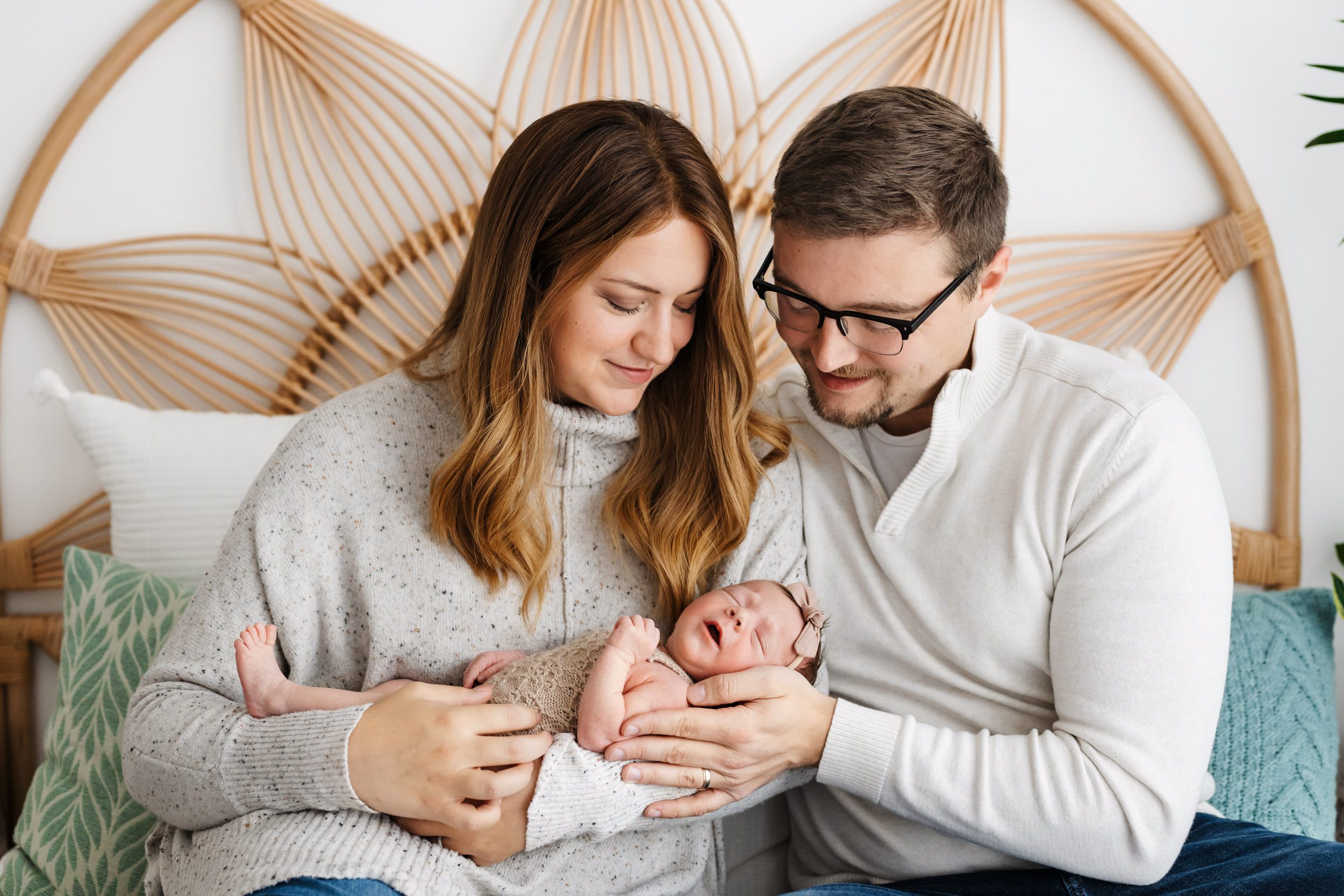 new parents sitting on a bed and smiling down at their baby girl cradled in mom's arms during a newborn photos session