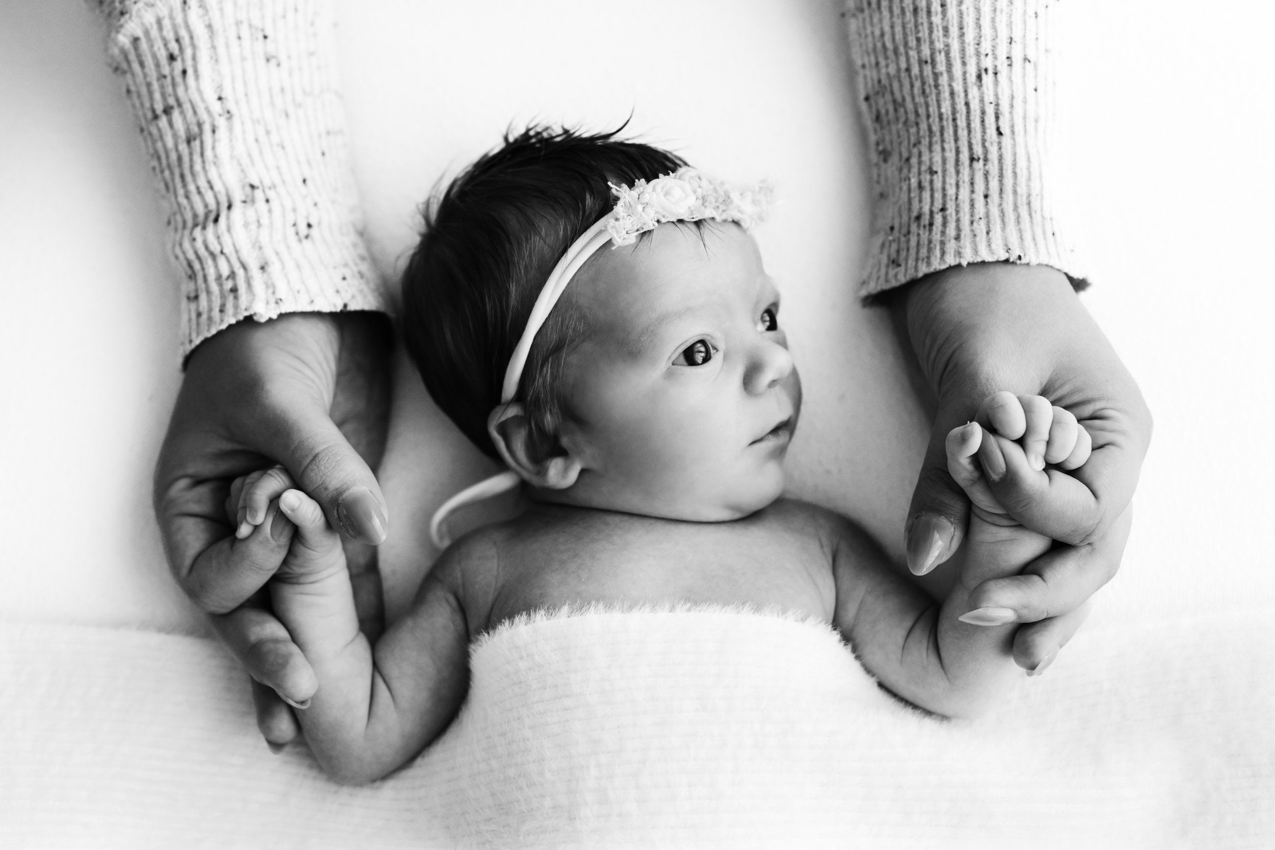 a black and white picture of a baby girl holding onto her mom's fingers and looking off to the right toward the window light with catch lights in her eyes during a newborn photos session
