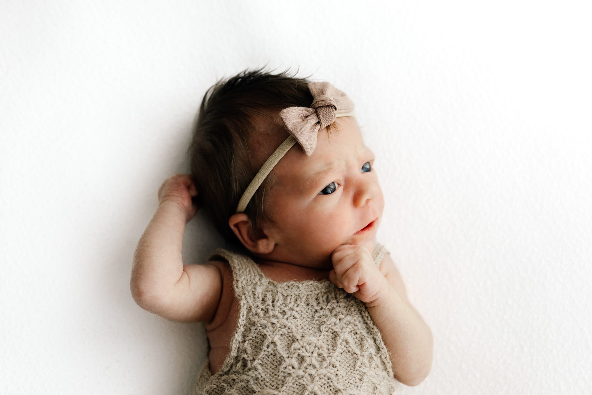 a baby girl wearing a textured, taupe colored knit romper laying on a white backdrop and touching her hand to her chin as she gazes off to the side during a newborn photography session