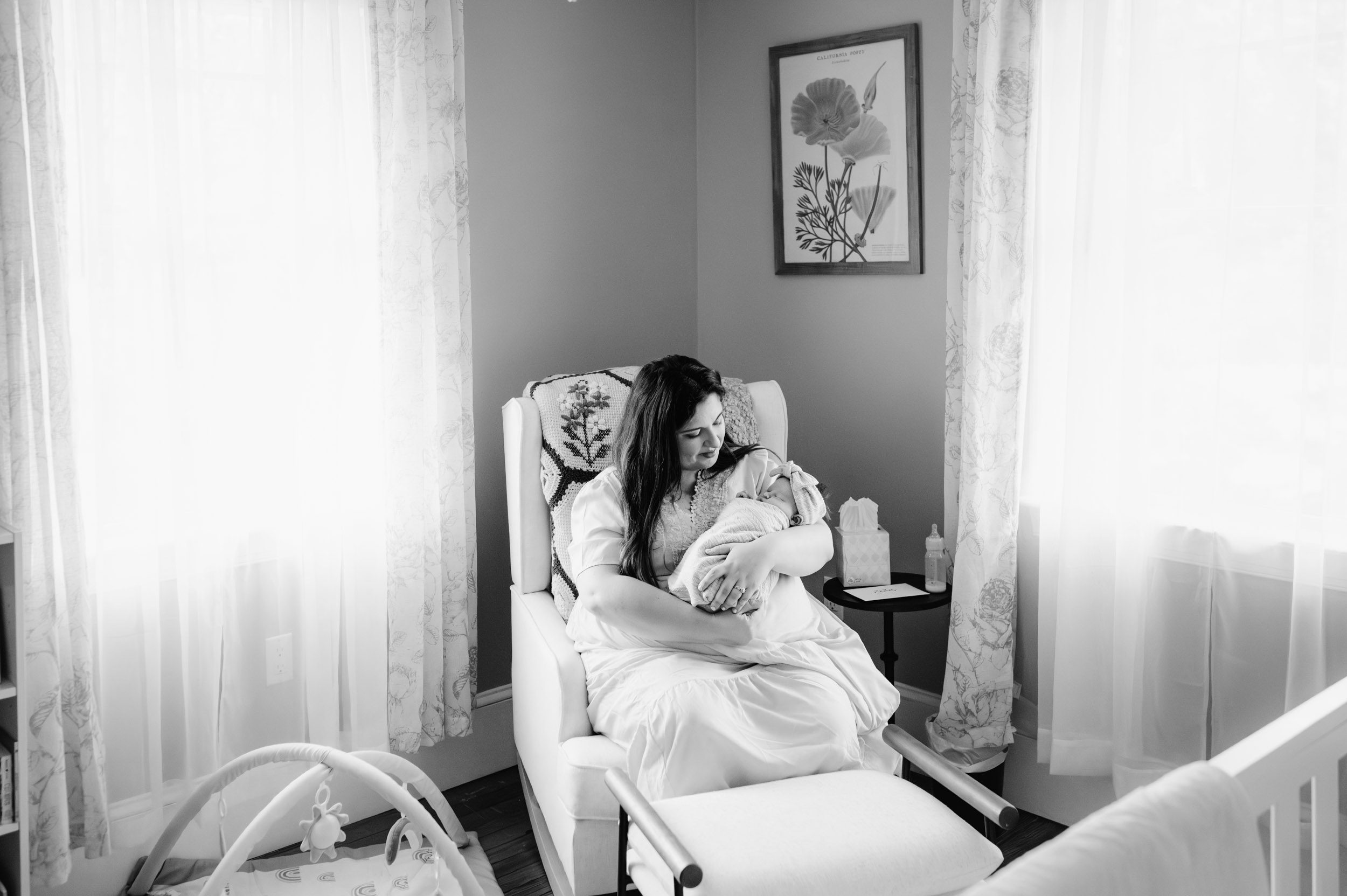 a black and white picture of a new mom sitting in a rocking chair in the baby's nursery and smiling down at her baby girl cradled in her arms during a Glenside newborn photoshoot