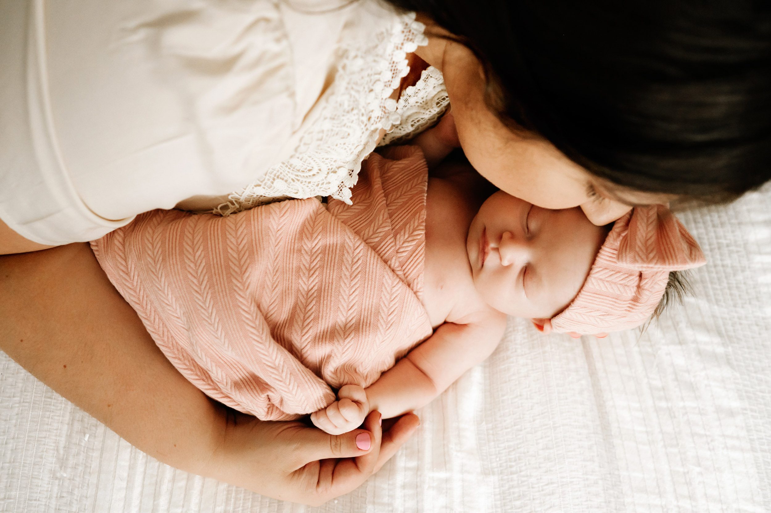 a picture taken from above of a baby girl laying on a bed wrapped in a pink swaddle blanket with her mom laying next to her and kissing her temple during a newborn photoshoot
