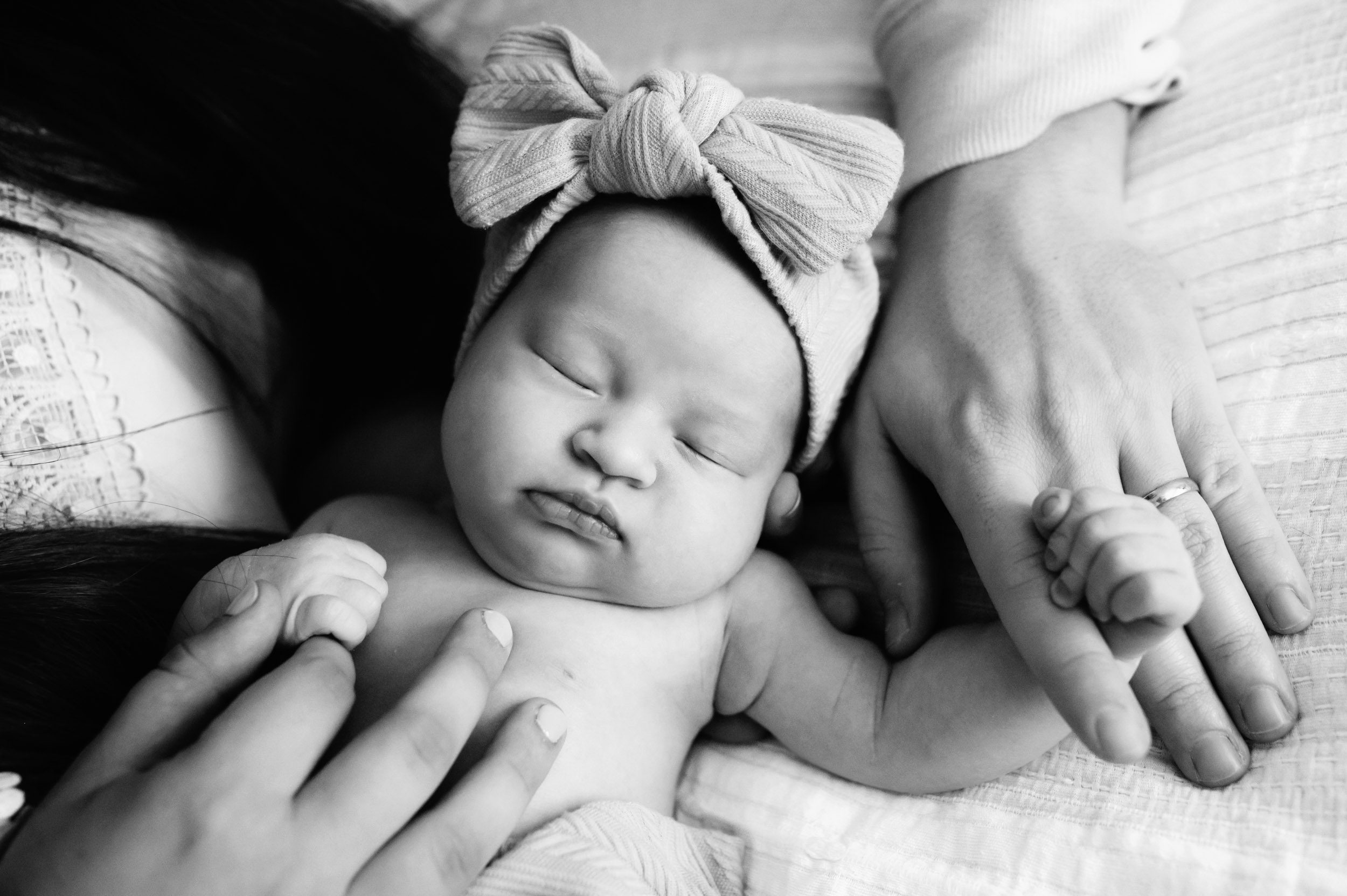 a black and white close up picture of a baby girl sleeping on a bed nestled up next to mom as she holds onto her parents' fingers with each of her hands