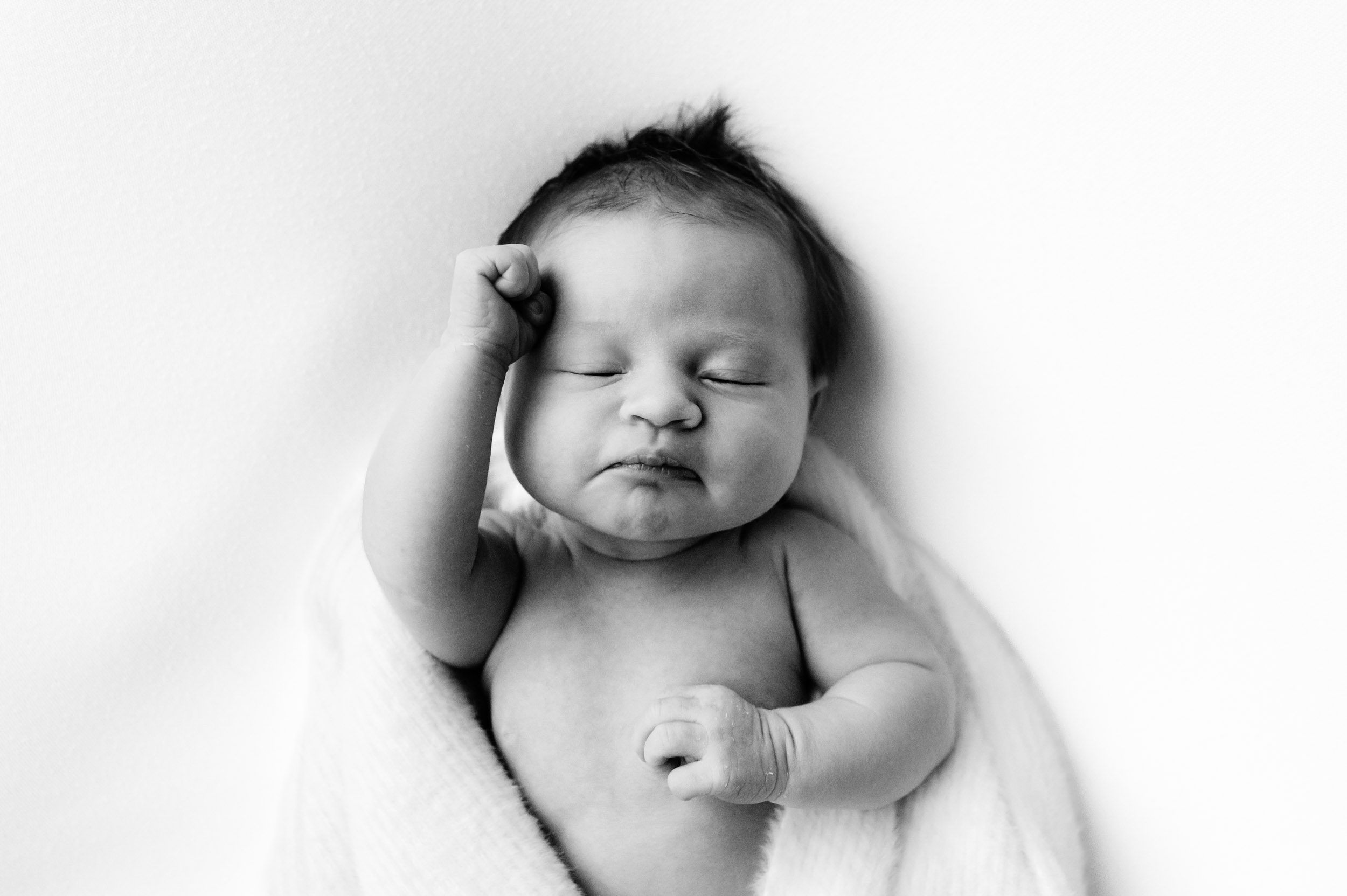 a black and white picture of a baby girl laying on a white backdrop with a fuzzy white blanket wrapped around her touching her hand to her forehead